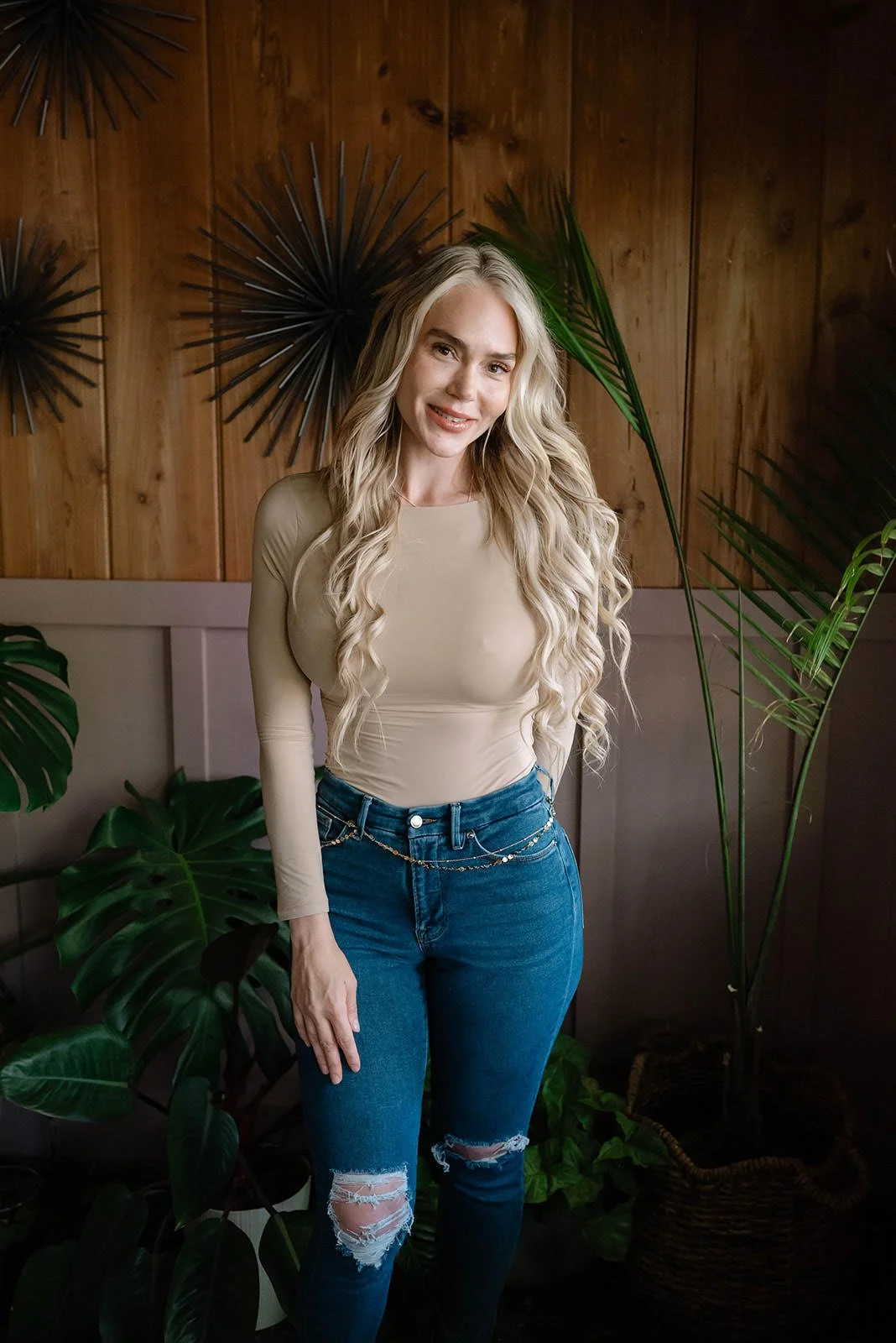 A woman with long, wavy blonde hair wearing a beige long-sleeve top and ripped blue jeans standing in front of a wooden wall decorated with black spiky wall art and surrounded by green plants.