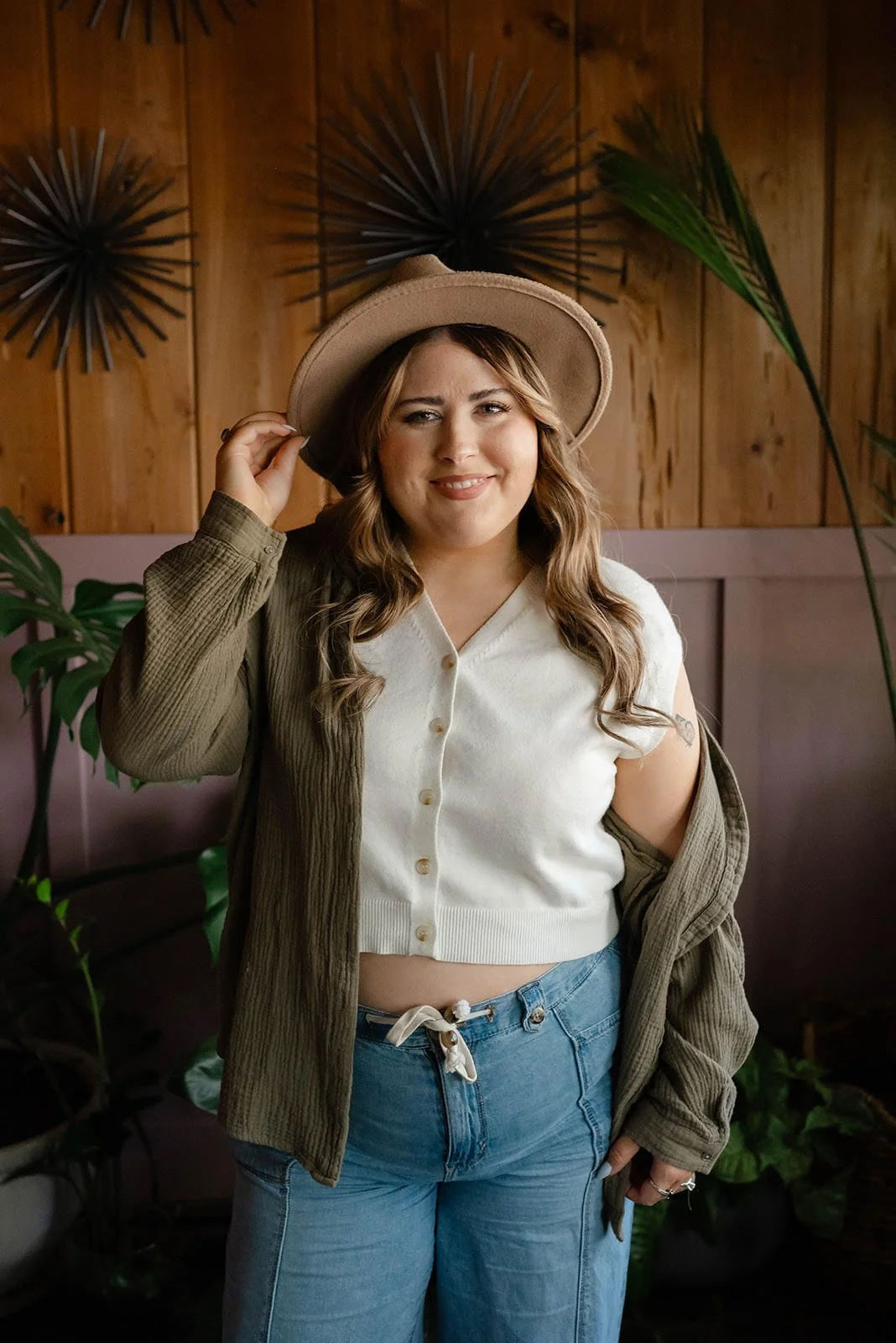 A young woman with long wavy hair wearing a beige hat, white sleeveless button-up top, olive green jacket, and blue jeans standing indoors with a wooden wall and plants in the background.