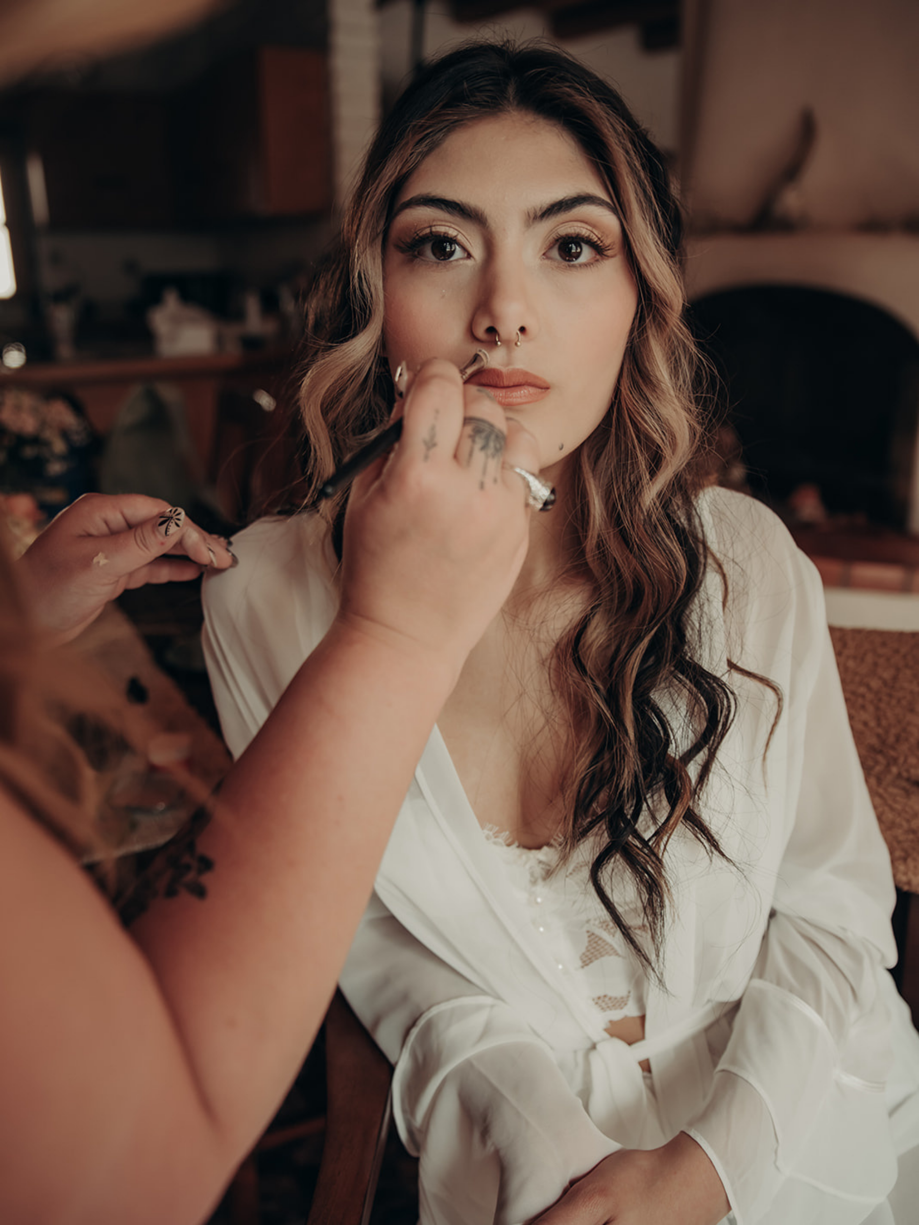 A woman with long, wavy hair sitting in a chair, having makeup applied to her lips, in a cozy indoor setting.