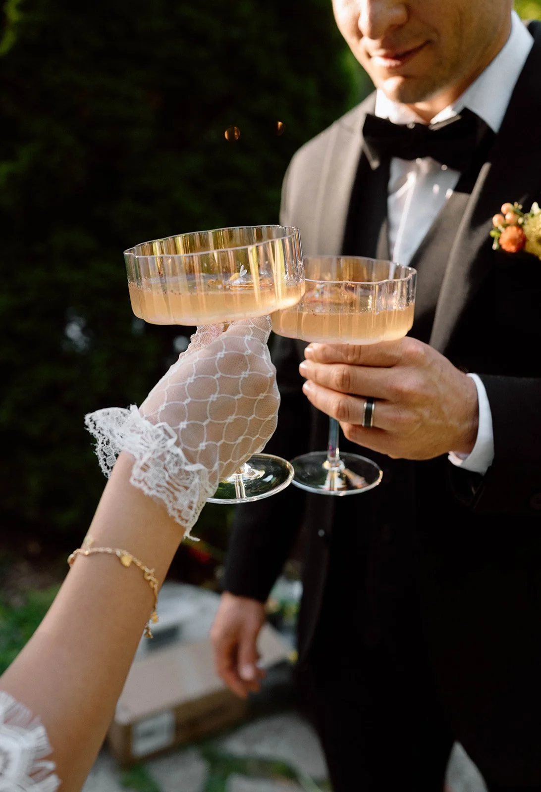 Person in a wedding tuxedo and another in a lace glove clinking champagne glasses at an outdoor celebration.