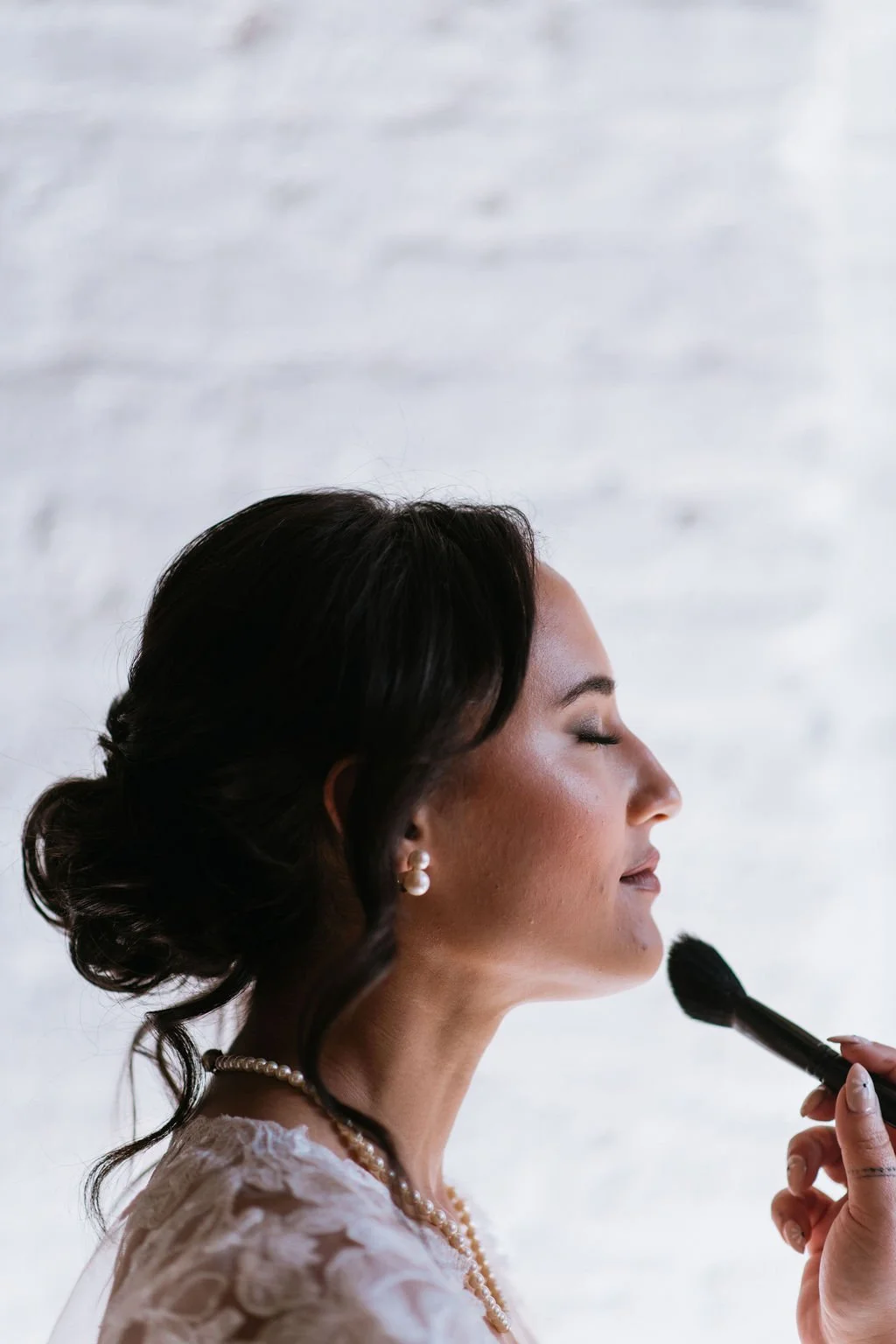 A woman with dark hair in an elegant updo, wearing pearl earrings and a pearl necklace, is getting her makeup done with a makeup brush.