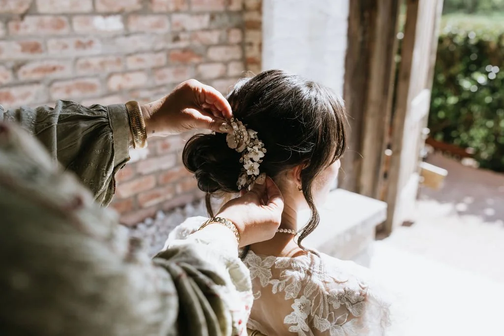 A woman is having her hair styled with a floral hair accessory outside near a brick wall. Someone is adjusting the woman's hair, which is up in a bun with loose curls.