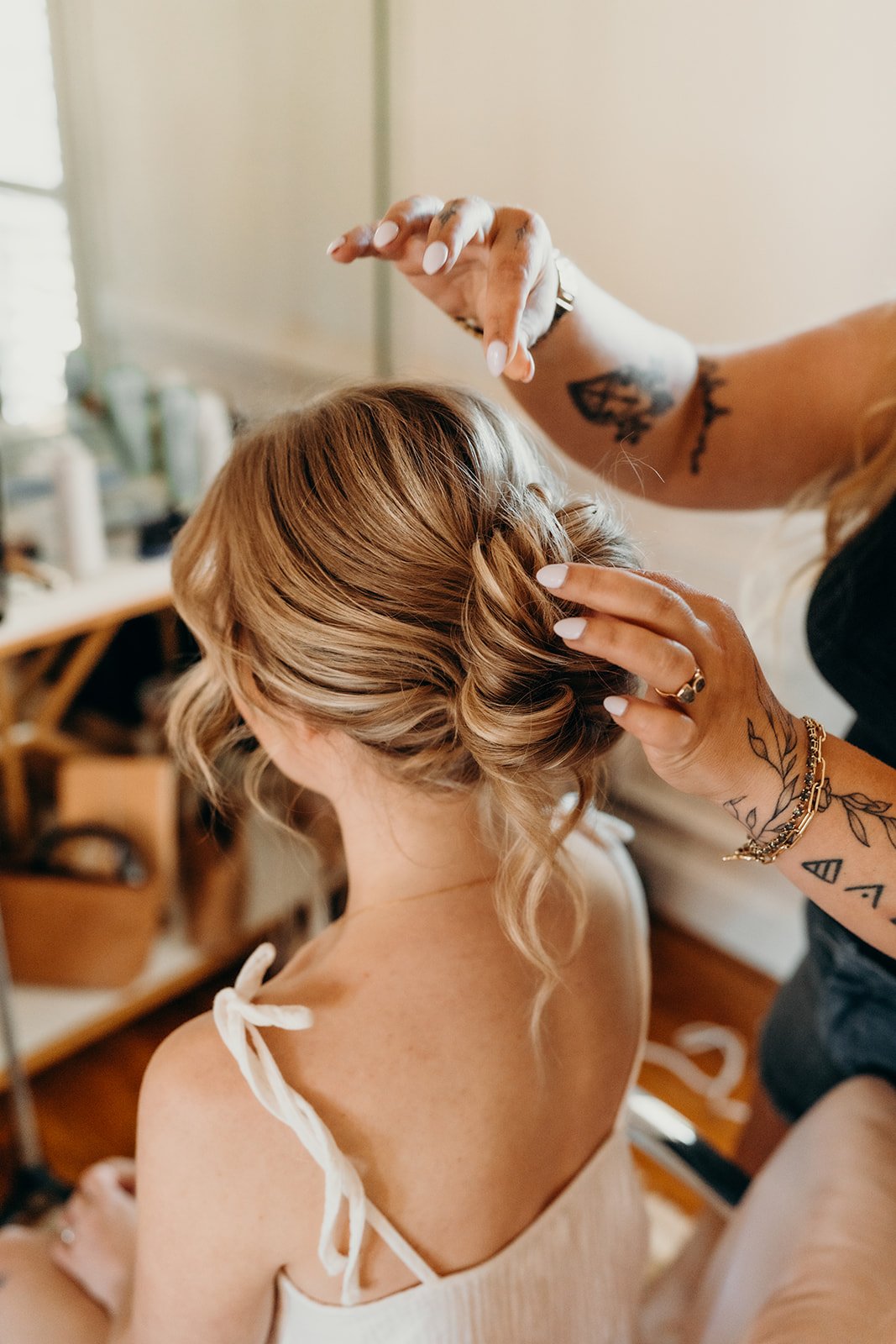 A woman with styled blonde hair getting her hair done by a hairstylist with tattoos and jewelry in a salon.