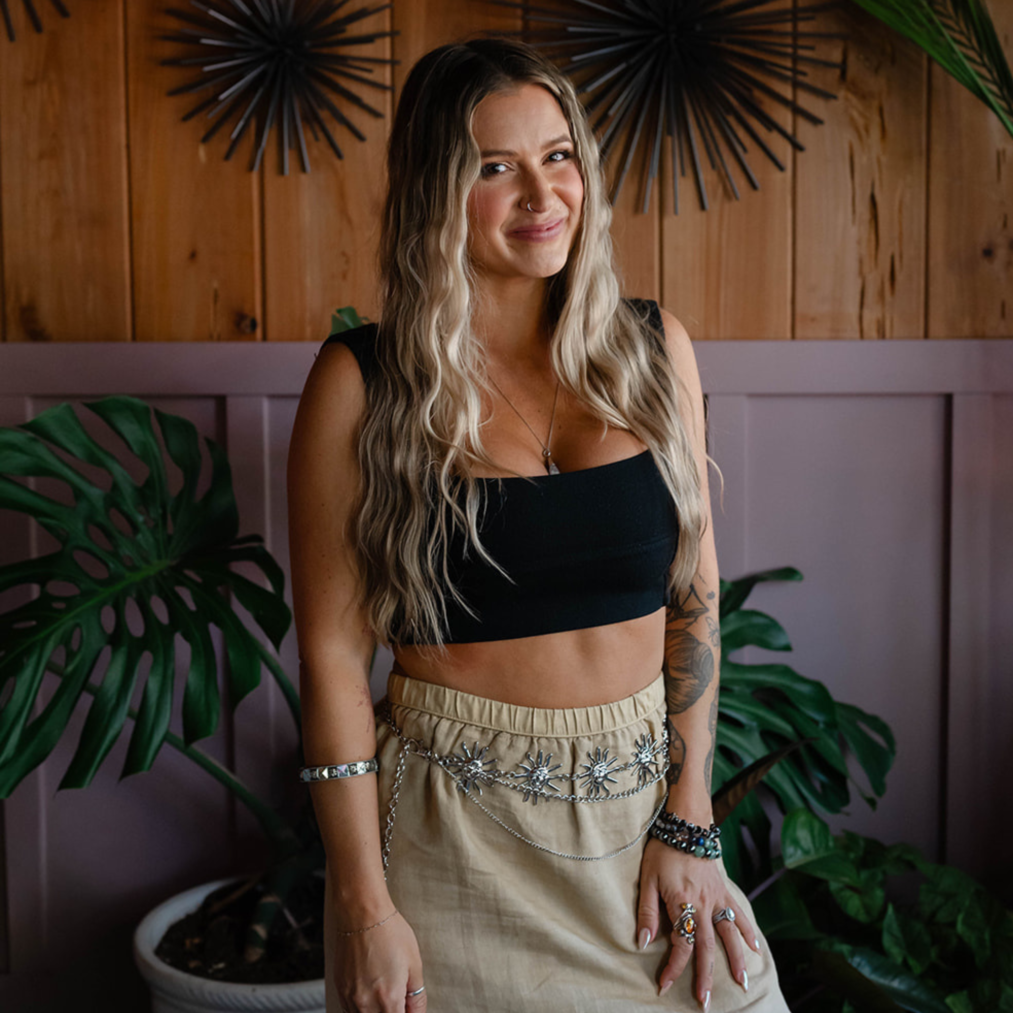 A woman with long, wavy blond hair, wearing rings, bracelets, and a nose piercing, is standing indoors in front of a wooden wall adorned with decorative black metal art and large green plants.
