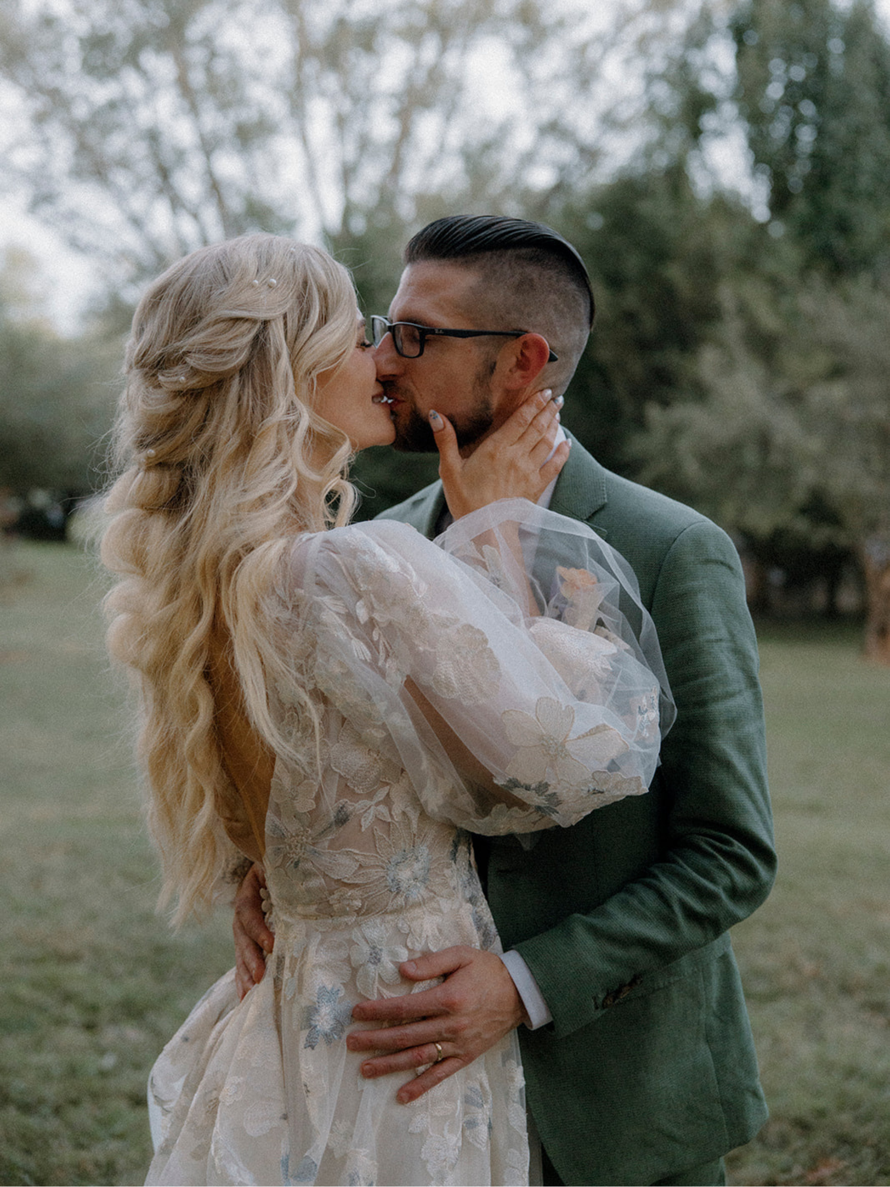 A couple kissing outdoors, woman in a lace dress and man in a suit, surrounded by trees.