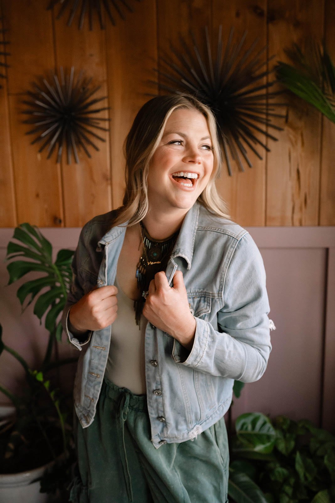 A woman with blonde hair smiling and looking to the side, holding her denim jacket, standing indoors with wooden wall decor and green plants in the background.