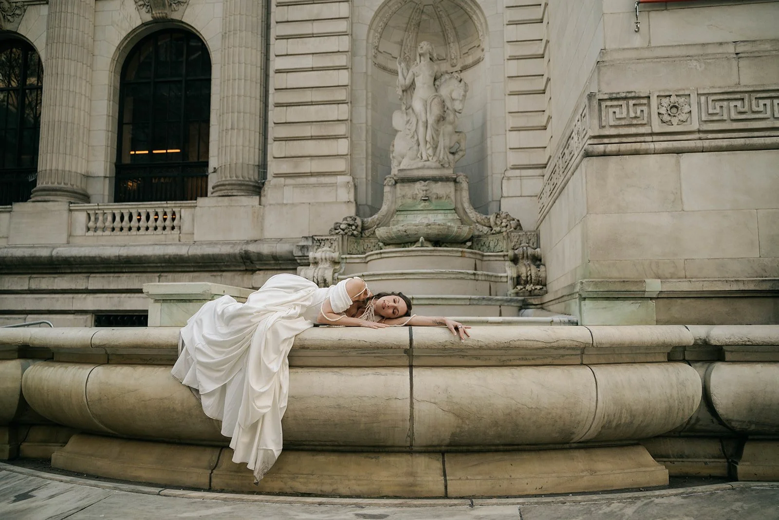 Woman in a white dress lying on a stone fountain in front of a historic building with classical columns and a statue of a woman on horseback.