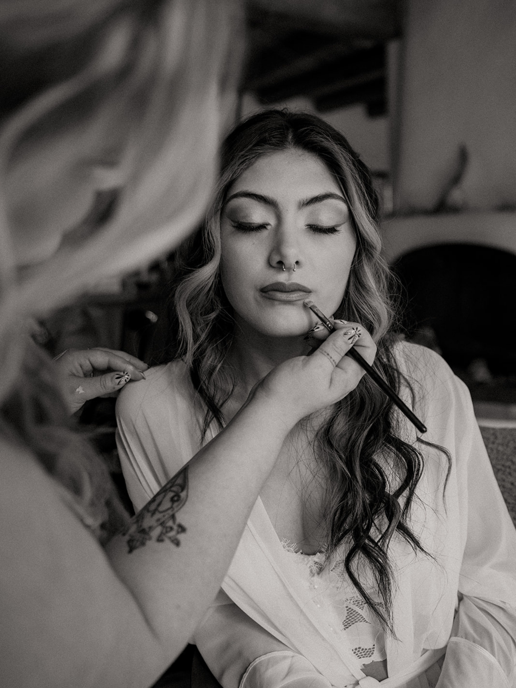 A young woman with closed eyes, long wavy hair, and a septum piercing has makeup applied by a makeup artist using a brush. The makeup artist's tattooed arm and hand are visible. The woman is wearing a light-colored blouse with lace details.