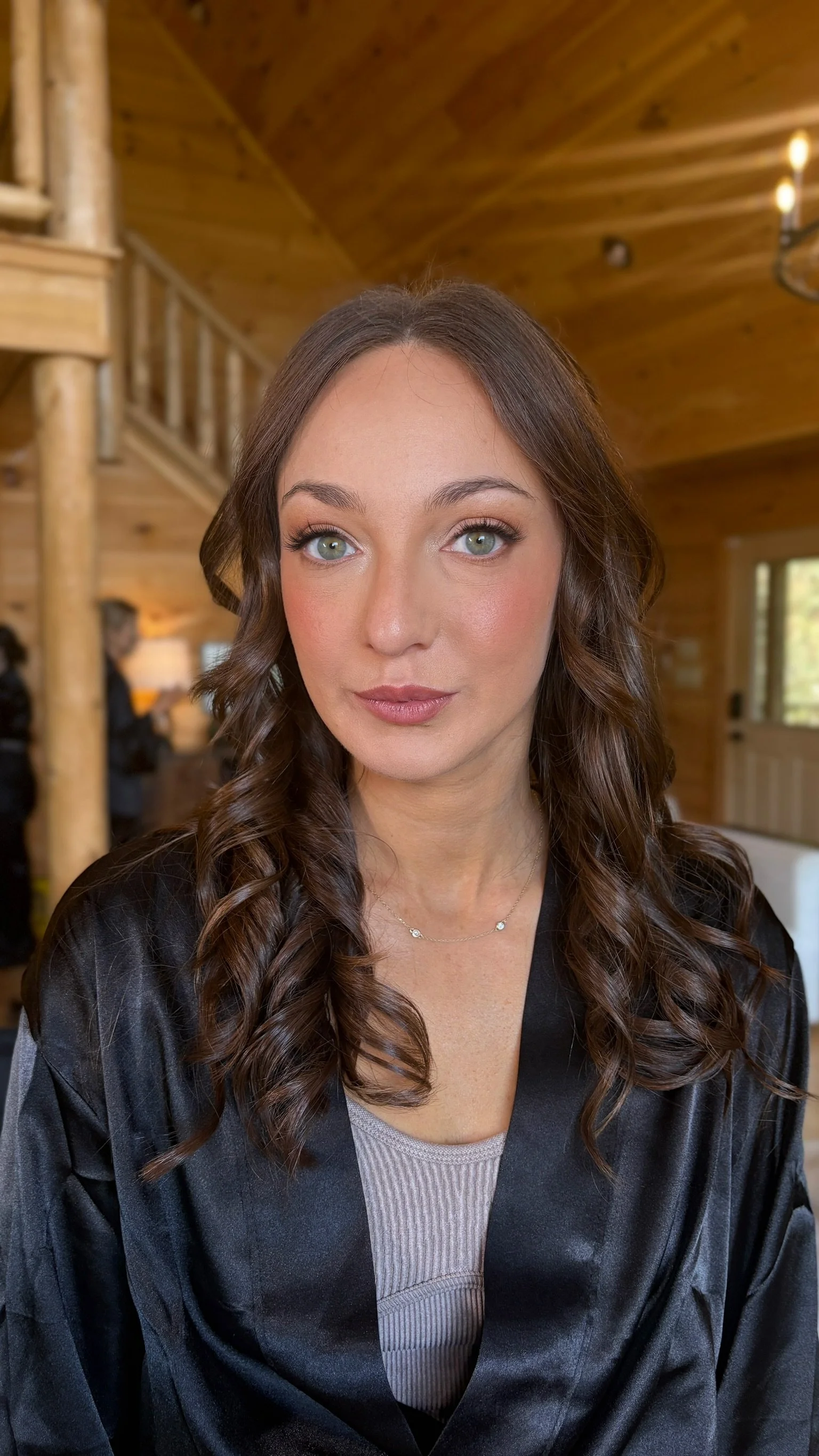 A woman with long, curly brown hair, wearing a black satin robe, a light gray ribbed top, and a delicate necklace, seated inside a wooden cabin with a staircase and chandelier in the background.