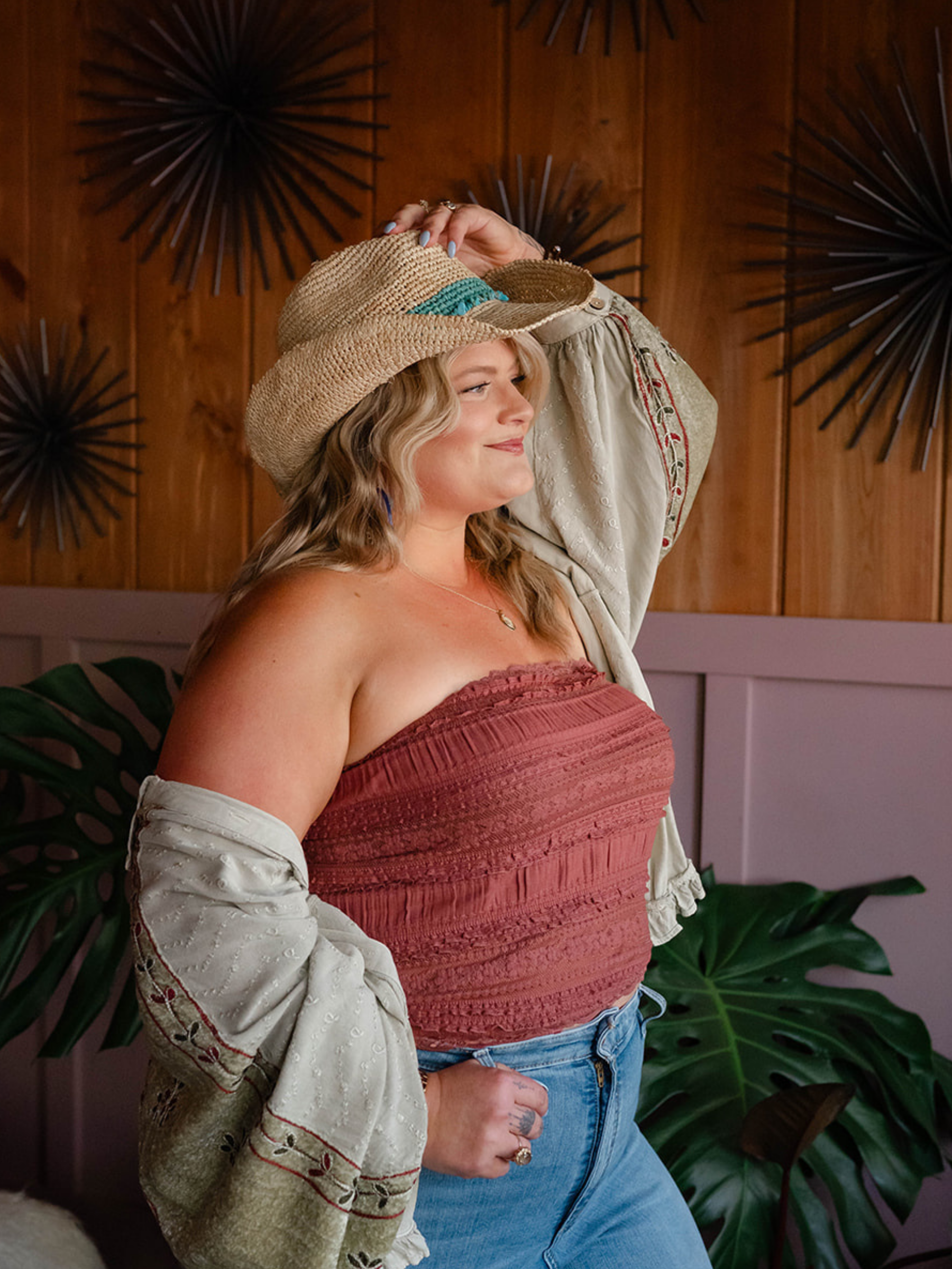 A woman in a red strapless top and jeans stands next to a large green plant, wearing a beige embroidered jacket and a wide-brimmed straw hat in front of a wooden wall with black metal decorative art.