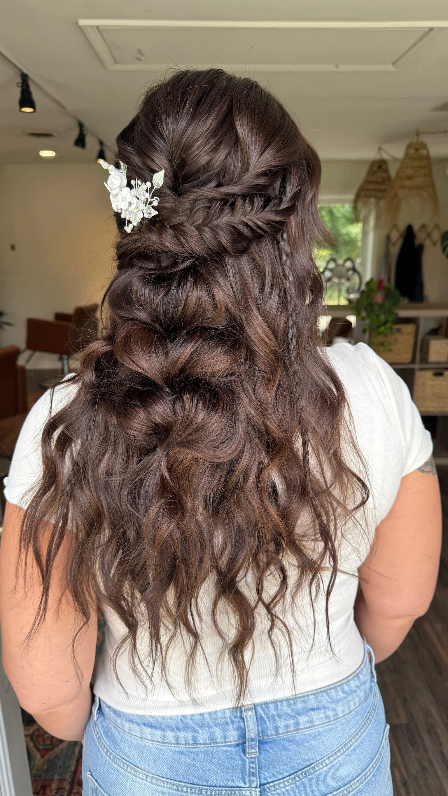 Woman with long, wavy brown hair styled with a twisted braid and decorated with a white floral hair accessory, standing indoors.