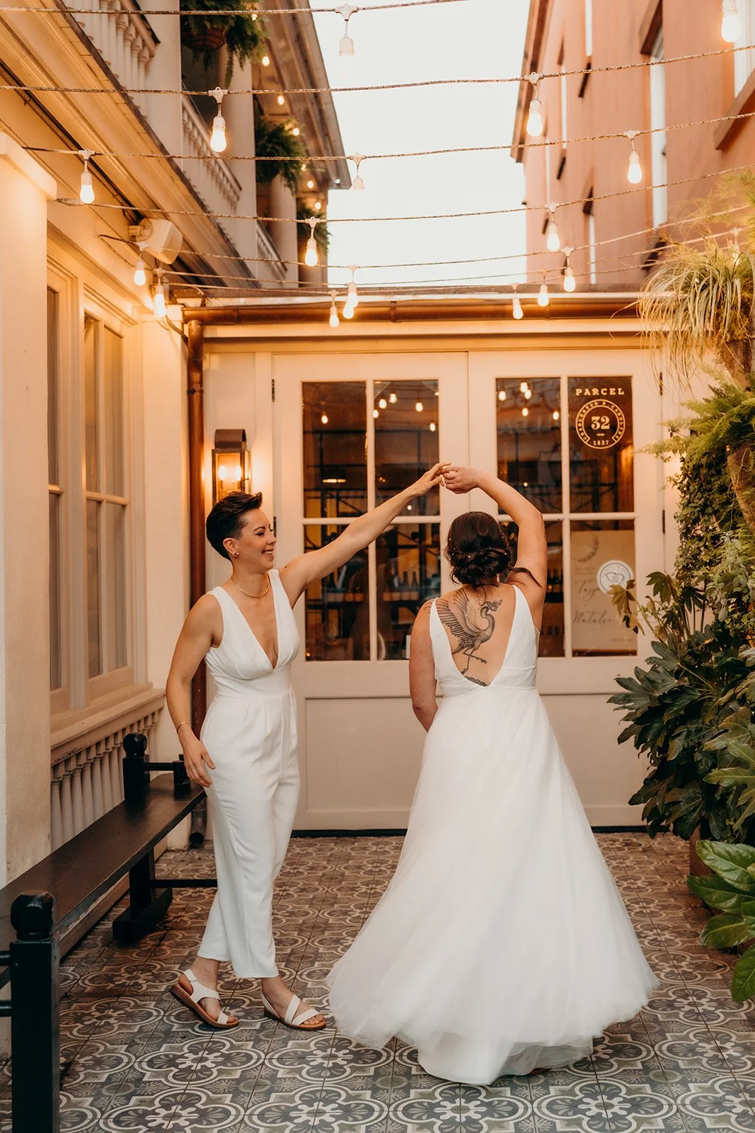 Two women in white dresses dance together in a beautifully lit outdoor patio, with string lights overhead and lush greenery around.