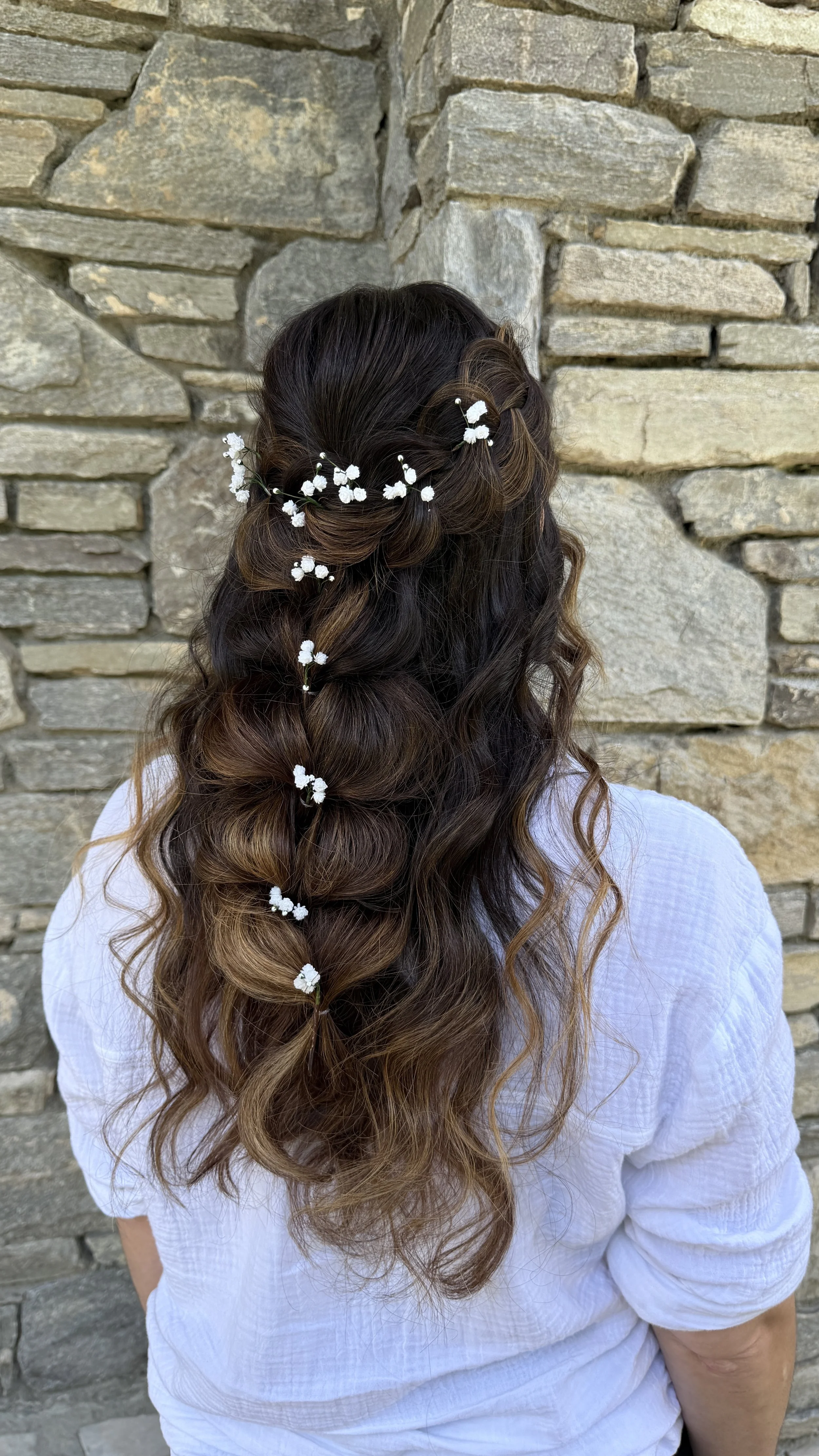 Woman with long, wavy hair styled in soft curls, decorated with small white flowers, standing against a stone wall.