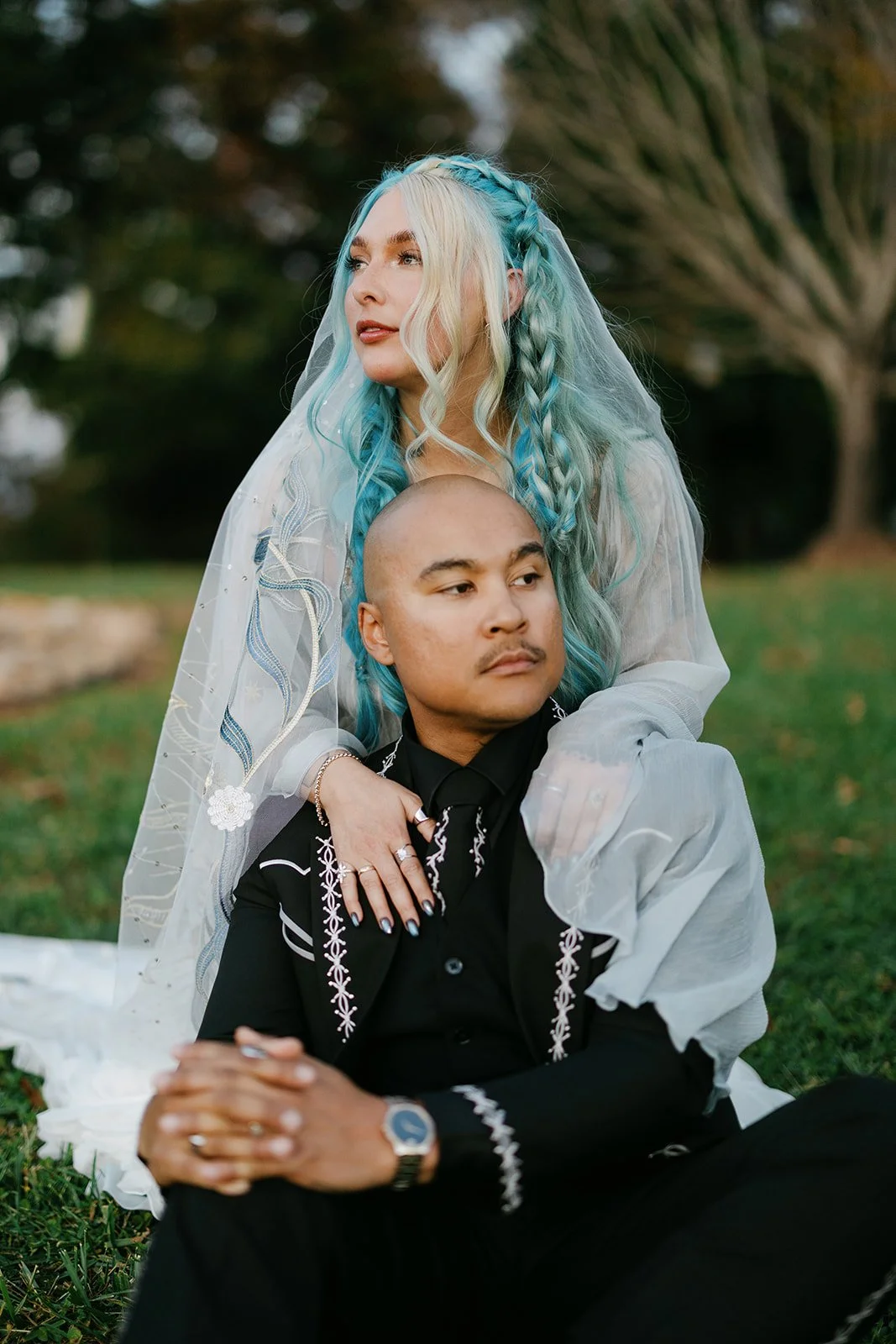 A woman with long, wavy blue and blonde hair, wearing a white sheer dress and veil, sits behind a seated man with a bald head, wearing a black embroidered shirt. They are outdoors on grass with trees in the background.