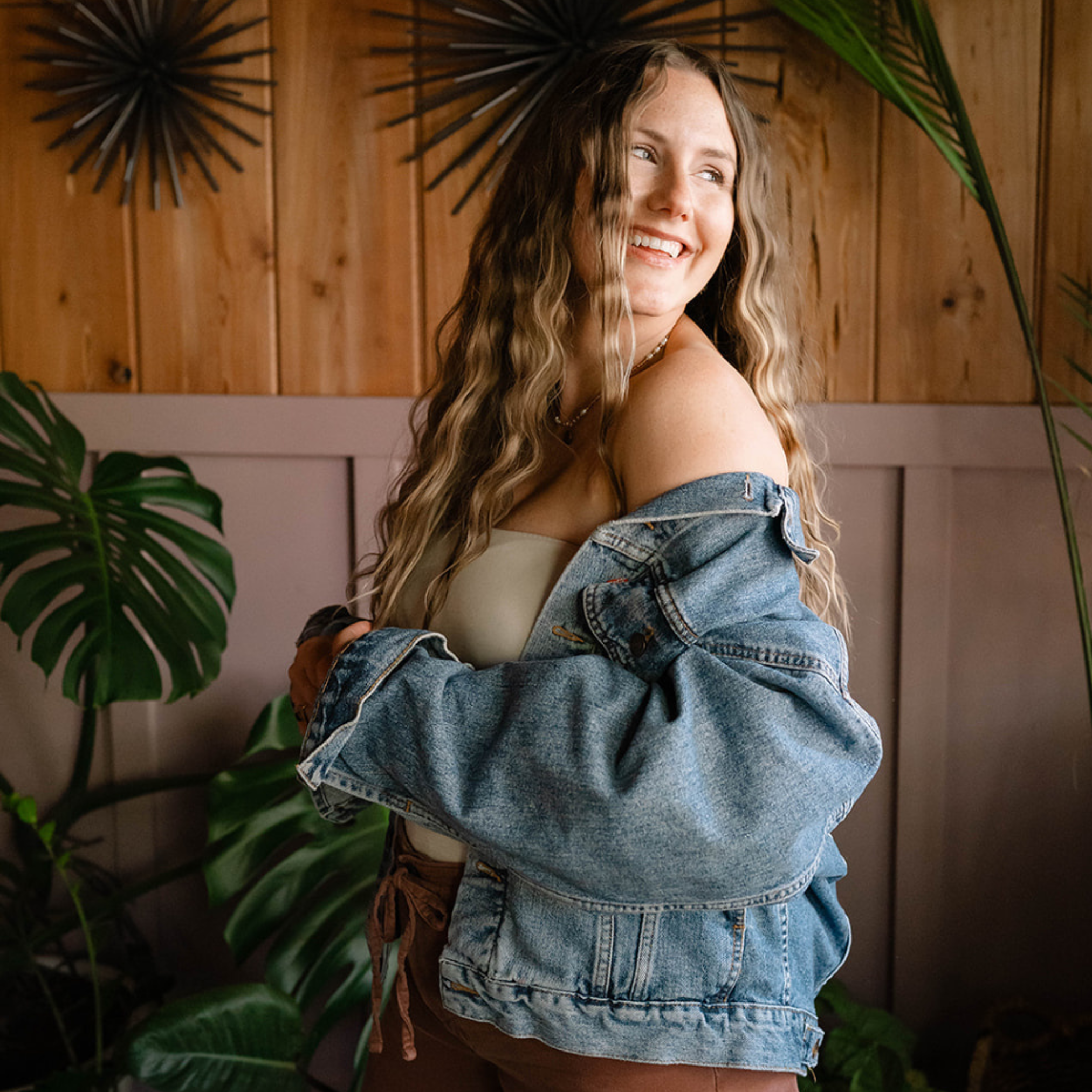 A woman with long wavy hair smiling in front of a wooden wall with decorative black sunburst wall art and green tropical plants.
