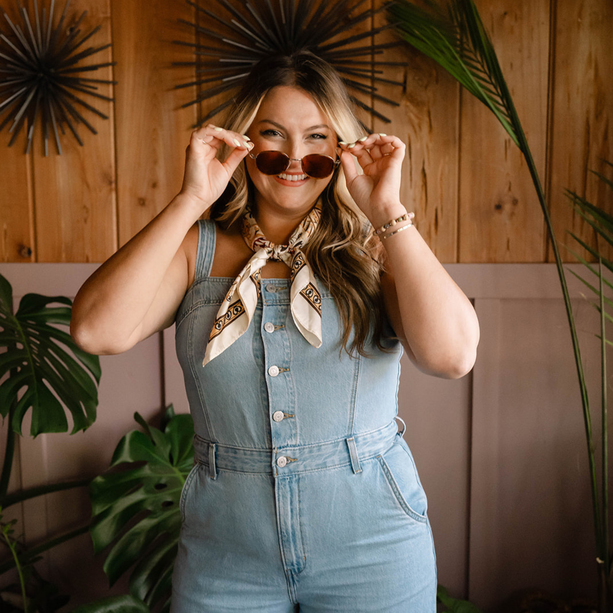 A woman smiling while holding sunglasses in front of her eyes, standing indoors with a wooden wall and large green plants in the background.