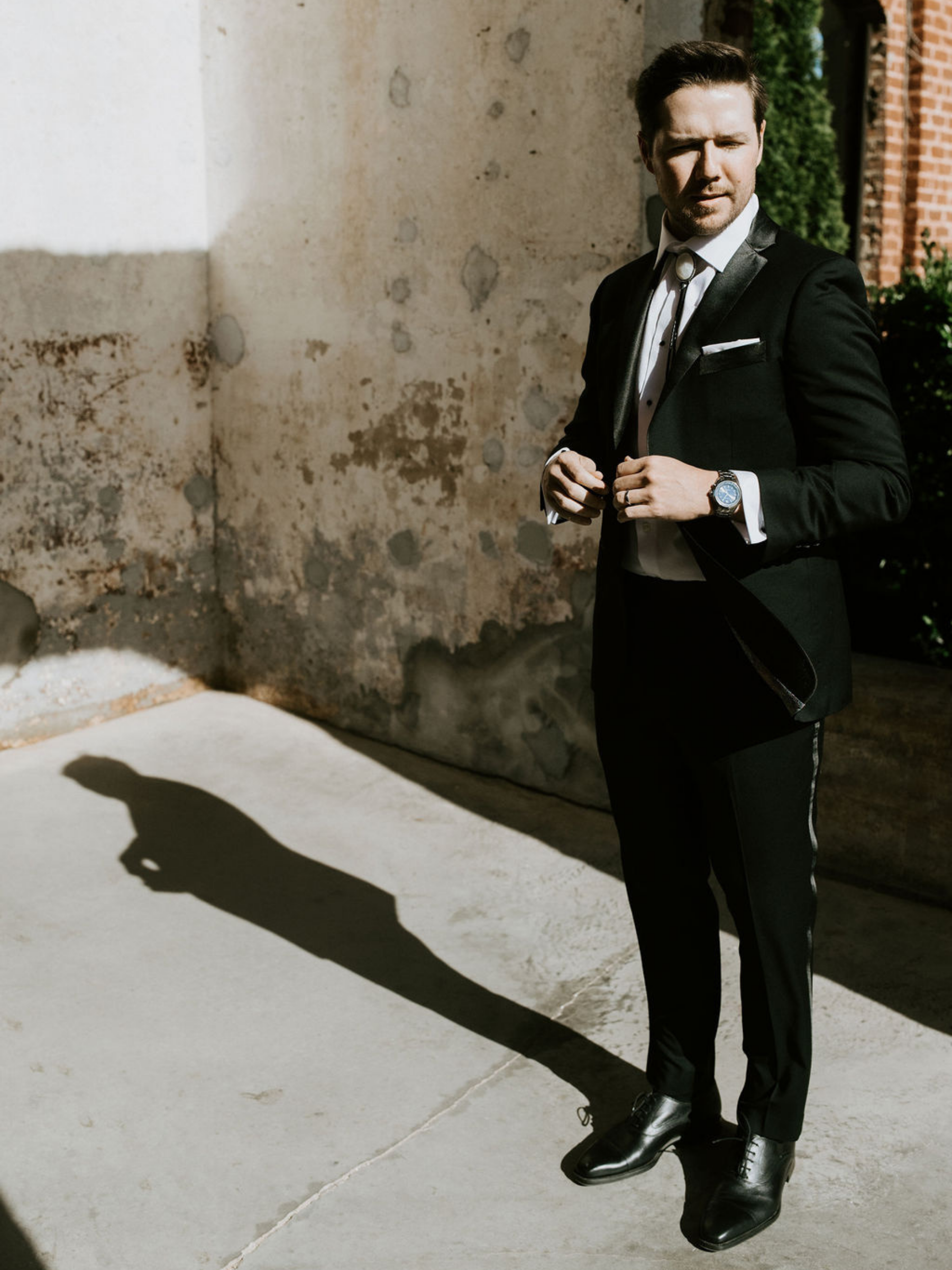 A man dressed in a black tuxedo with a white dress shirt, black shoes, and a watch, standing outdoors in sunlight, casting a shadow on the concrete ground.