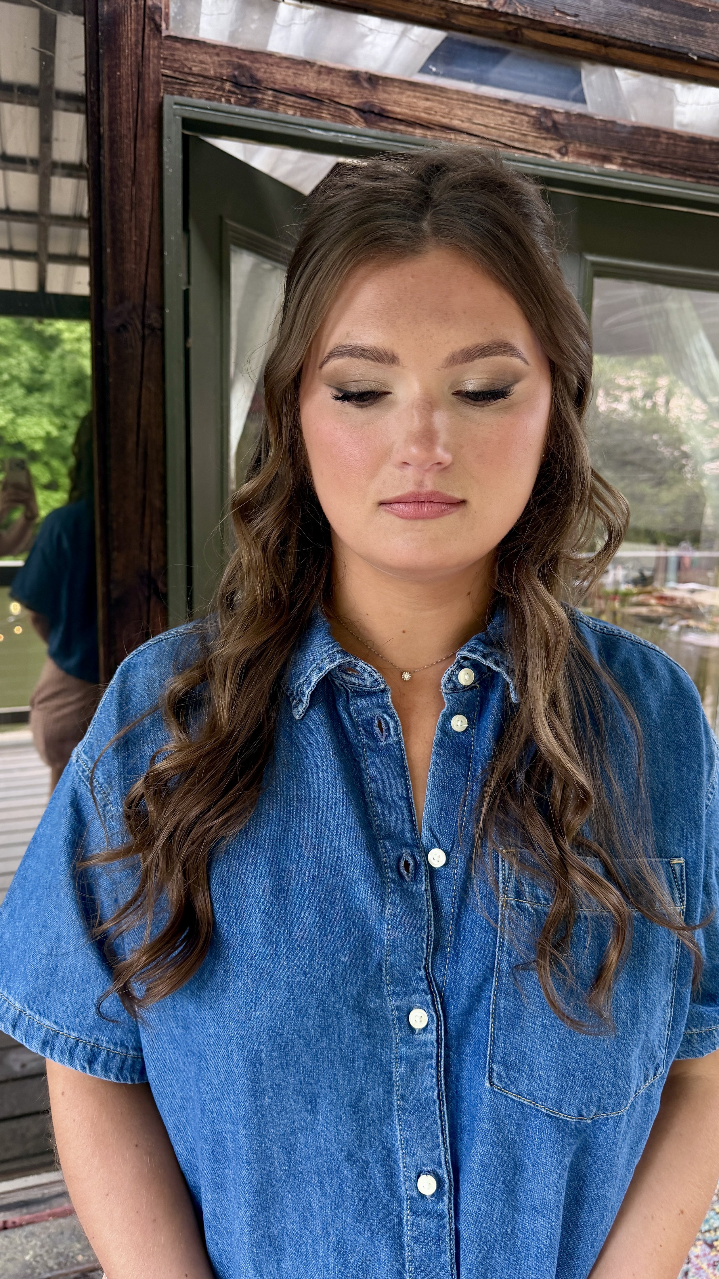 A young woman with long wavy hair wearing a blue denim shirt, standing outside near a wooden structure and a glass window.