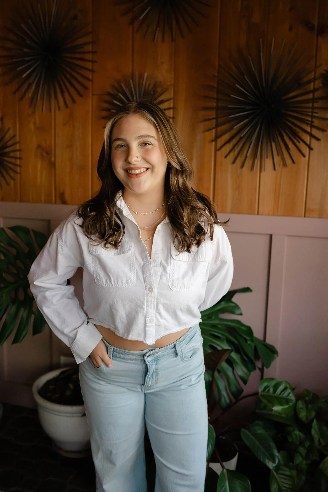 A young woman with dark brown, wavy hair, wearing a white cropped button-up shirt and light blue jeans, smiling and standing indoors in front of a wooden wall with decorative black metal wall art and green houseplants.
