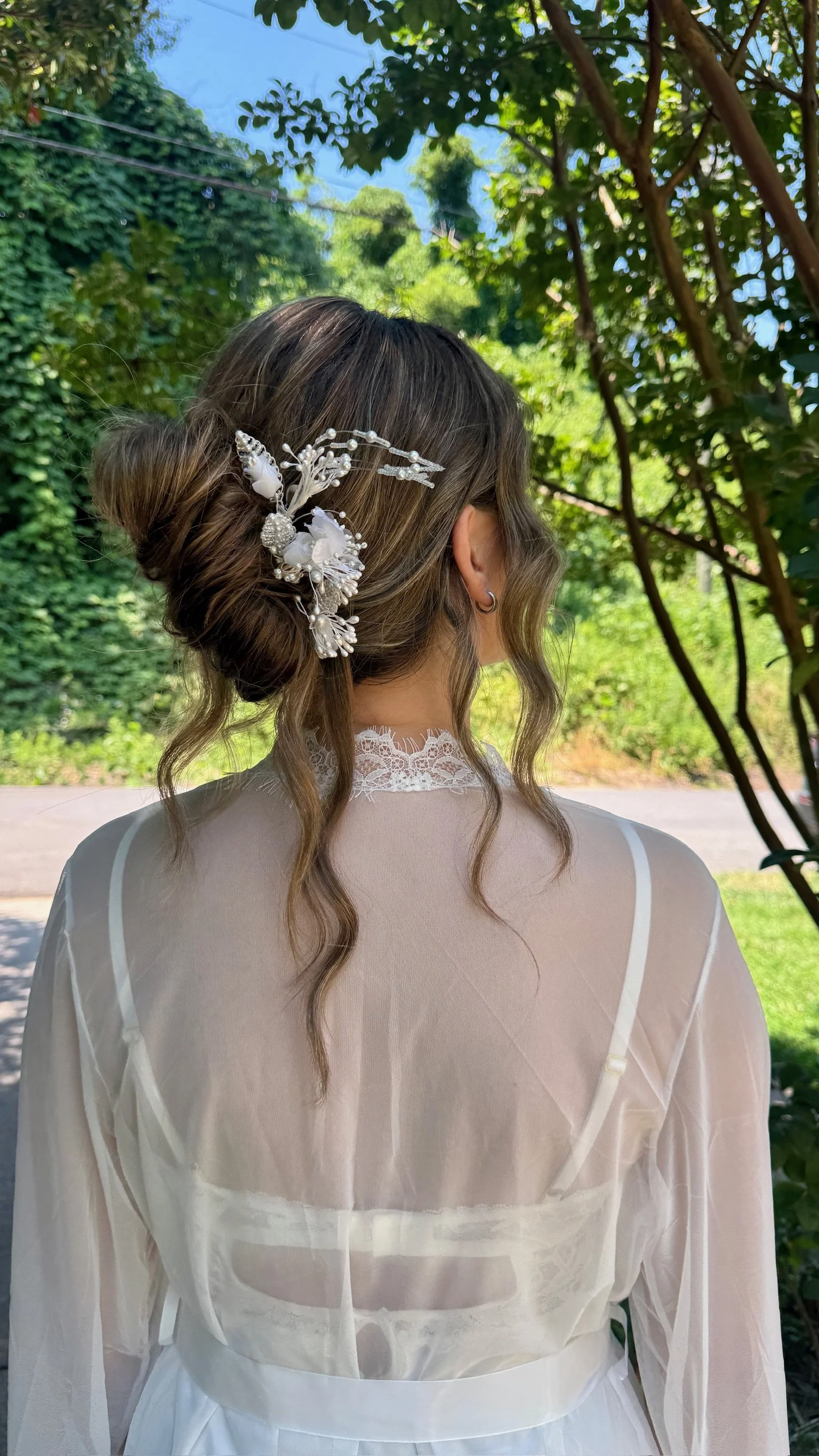 Woman with wavy hair styled in an updo, wearing a decorative hairpiece, seen from behind, outdoors with green trees and blue sky in the background. She is dressed in a sheer white garment with lace details.