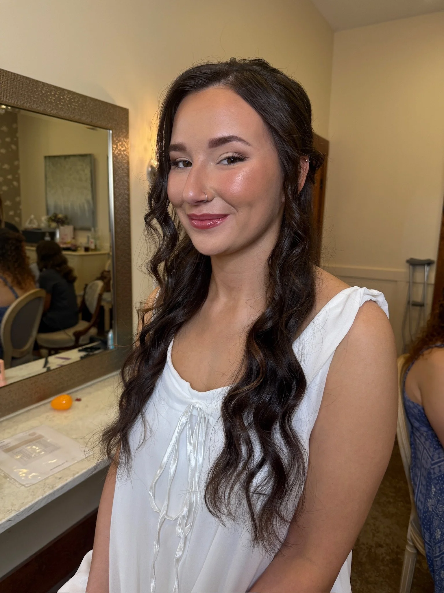 A woman with long dark curly hair styled in loose waves, wearing a white sleeveless top with a bow tie at the front, smiling at the camera inside a room with yellow walls and a mirror on the wall.