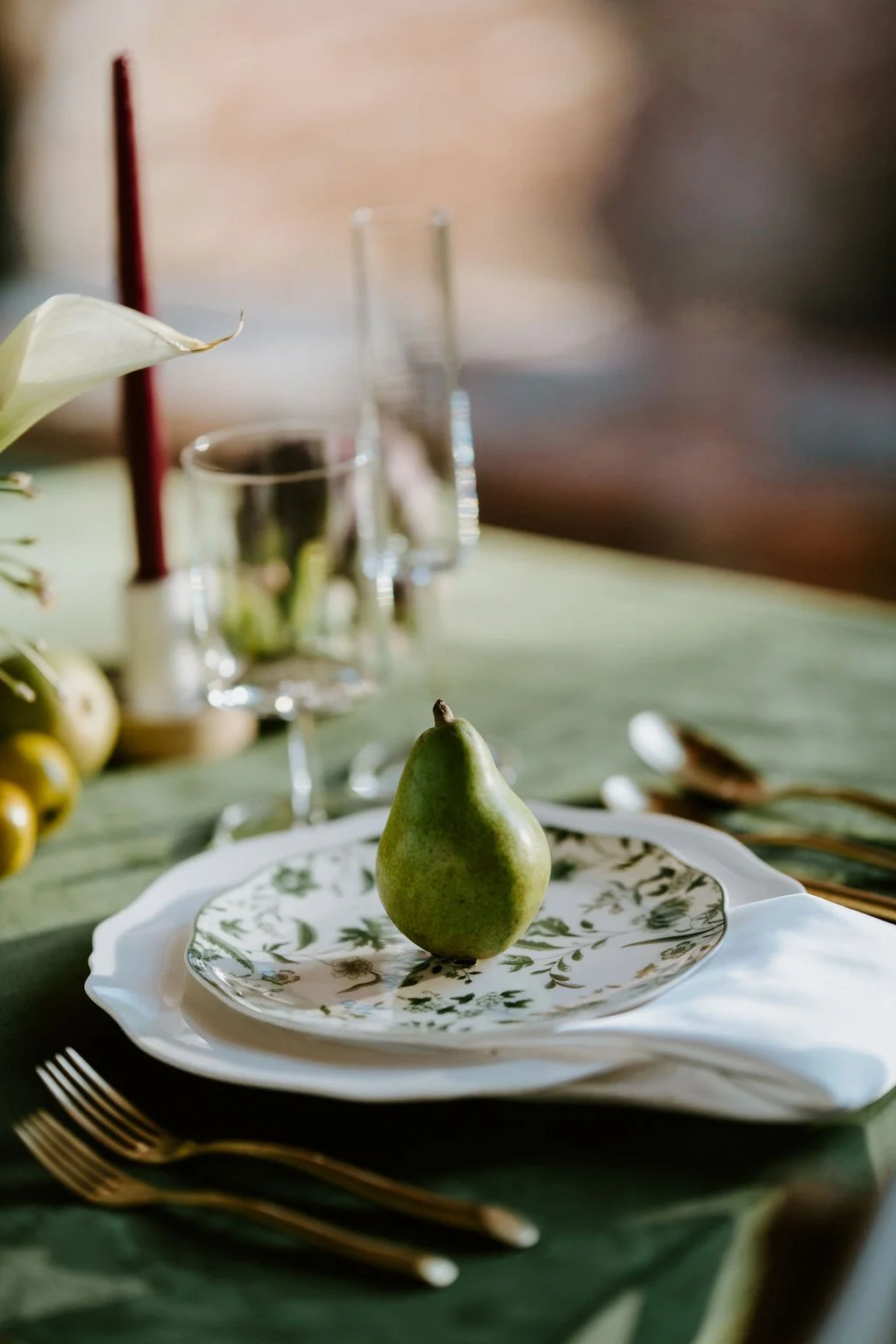 A decorated table with a green and white patterned plate holding a pear, set with gold utensils, and background items including glasses, a vase with flowers, and candles.