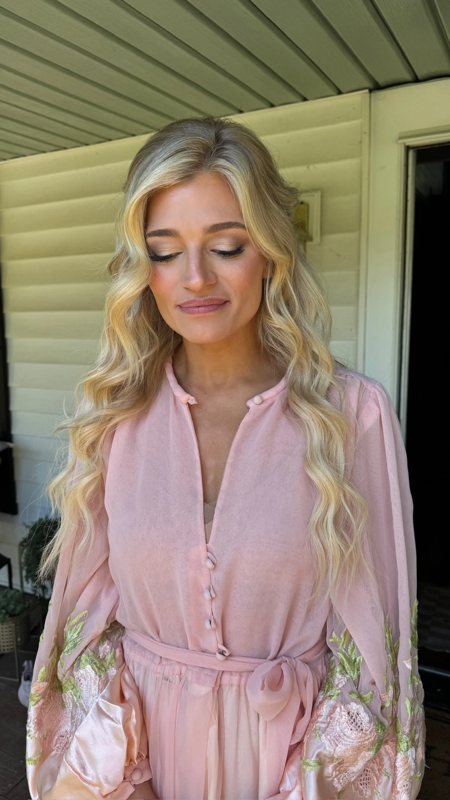 A woman with long blonde wavy hair, closed eyes, and wearing a pink dress with floral embroidery on the sleeves, standing outdoors in front of a beige house with a porch ceiling.