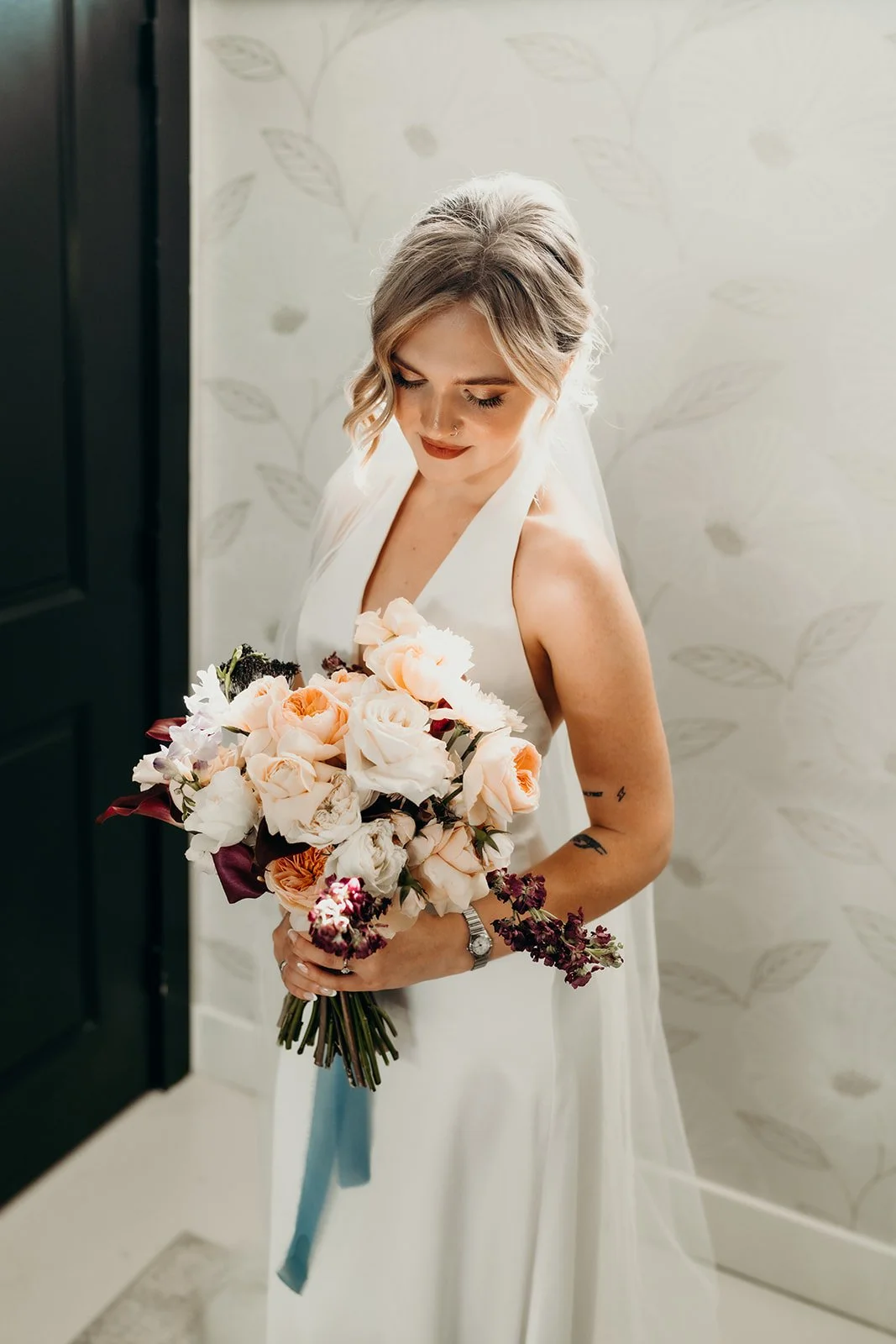A bride with blonde hair styled in loose waves holding a bouquet of pink, cream, and burgundy flowers, wearing a white sleeveless wedding dress, standing indoors near a wallpapered wall.