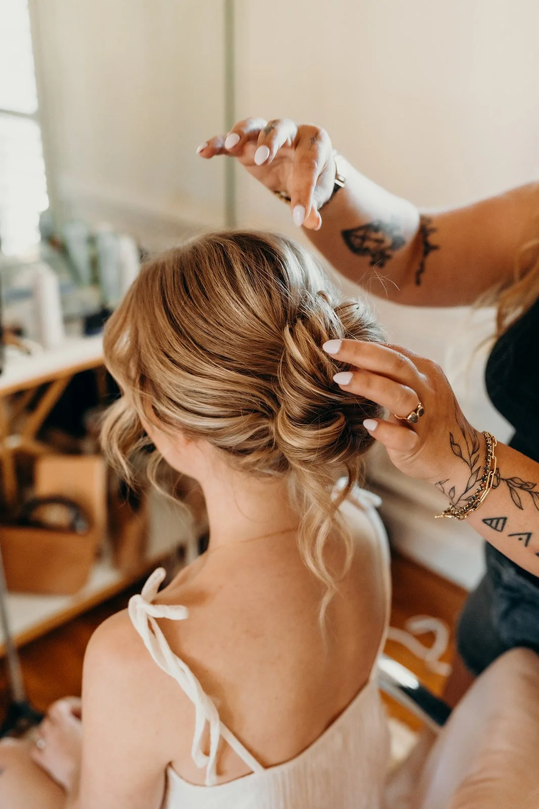 A hairstylist is styling a woman's light brown, wavy hair in an updo. The woman is wearing a white top with tie straps on her shoulders, and the stylist has tattoos on her arms. The background shows a salon or room with shelves and blurred objects.