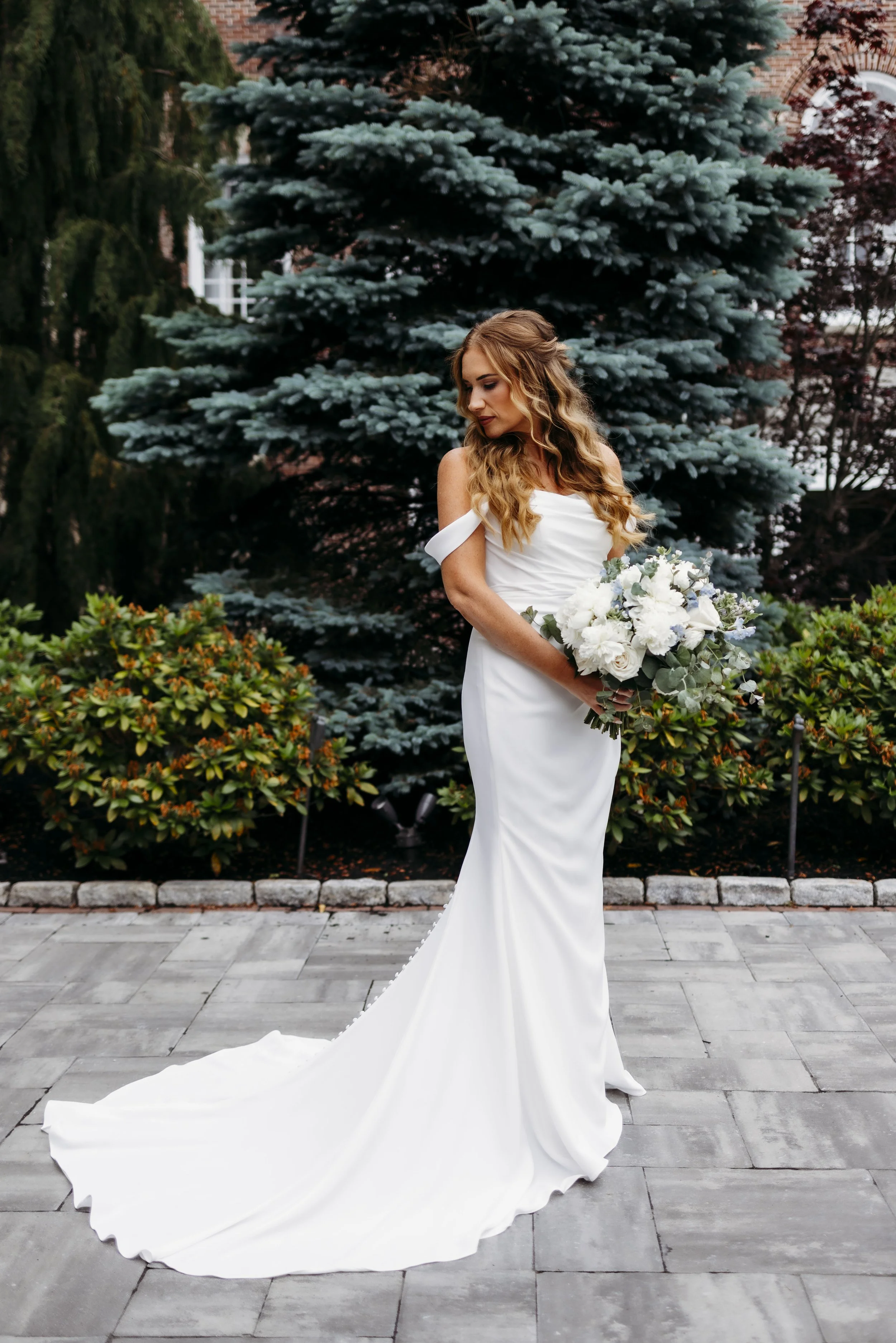 A woman in a white wedding dress holding a bouquet of white flowers, standing outdoors in front of green trees.