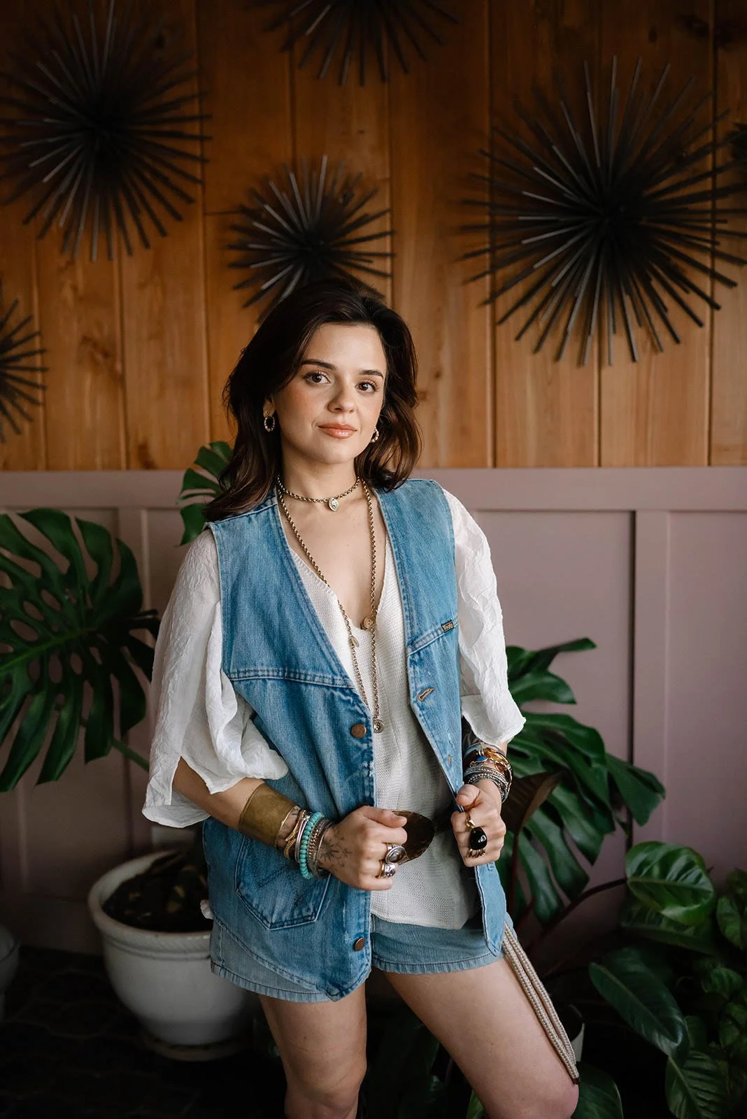 A woman standing indoors in front of a wooden wall with black starburst wall decor. She wears a denim vest over a white blouse, denim shorts, and multiple jewelry pieces, surrounded by green plants.