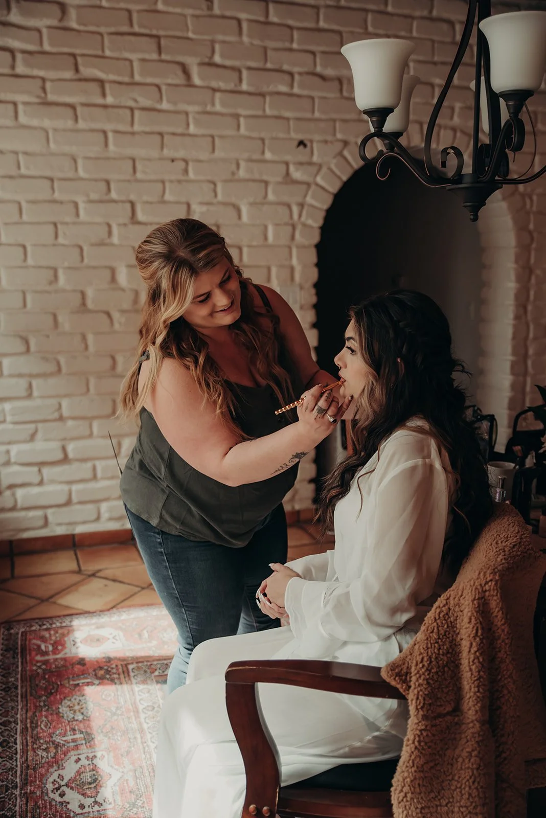 A makeup artist applying lipstick to a seated woman with long dark hair and a white robe, inside a room with a brick wall and chandelier.