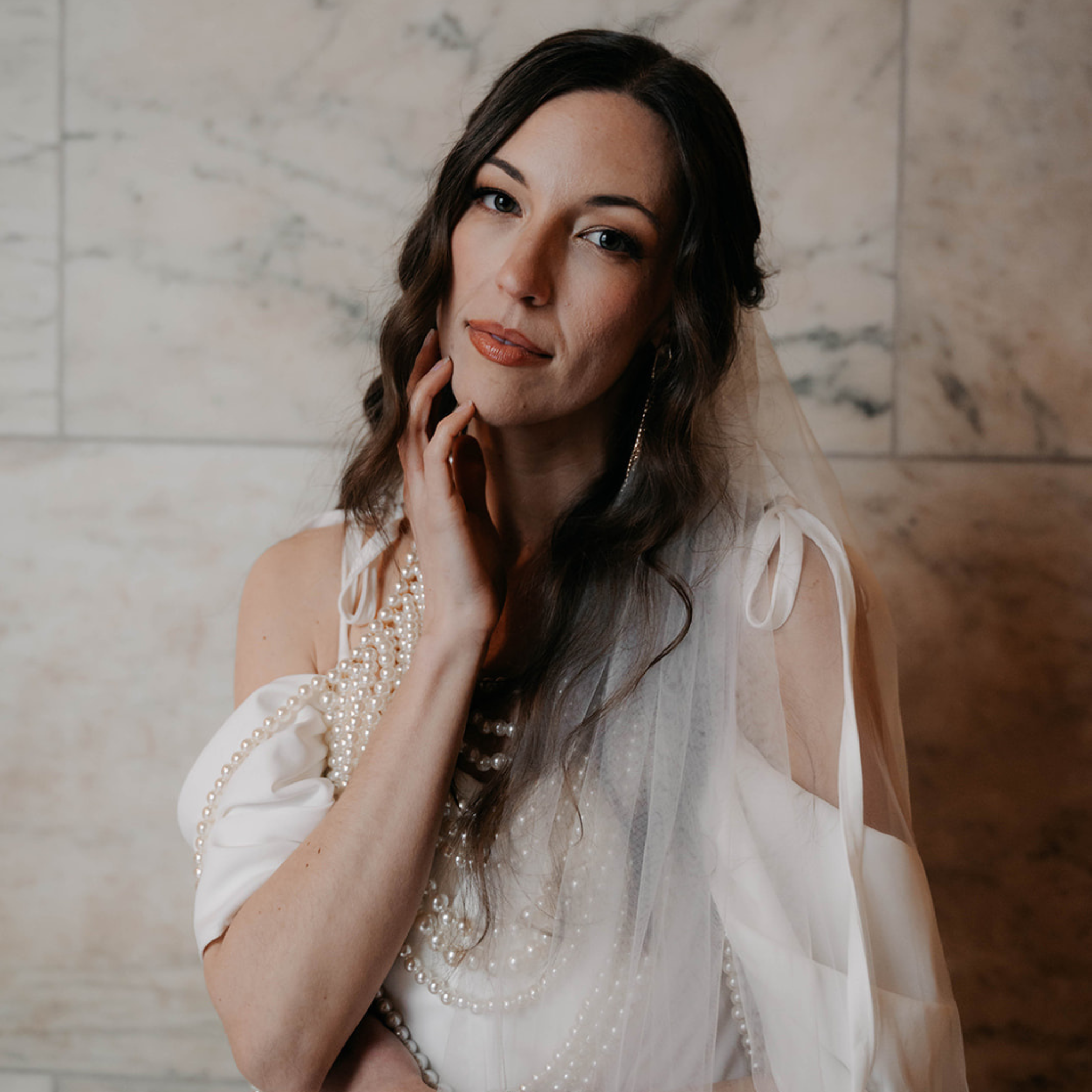 A woman with dark hair styled in loose waves, wearing a cream-colored dress adorned with pearl necklaces, touching her face with her right hand, standing in front of a marble wall.