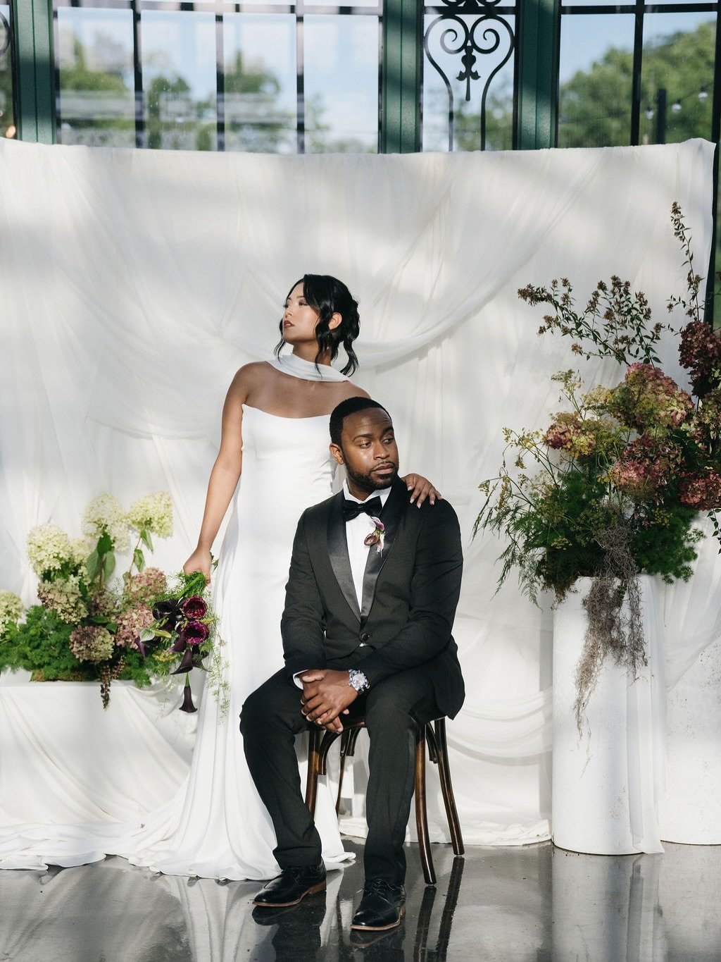 A woman in a white dress is standing behind a man in a black tuxedo, who is seated on a stool. They are in front of a white draped backdrop with floral arrangements on either side. The woman has dark, wavy hair and is looking away, while the man has 