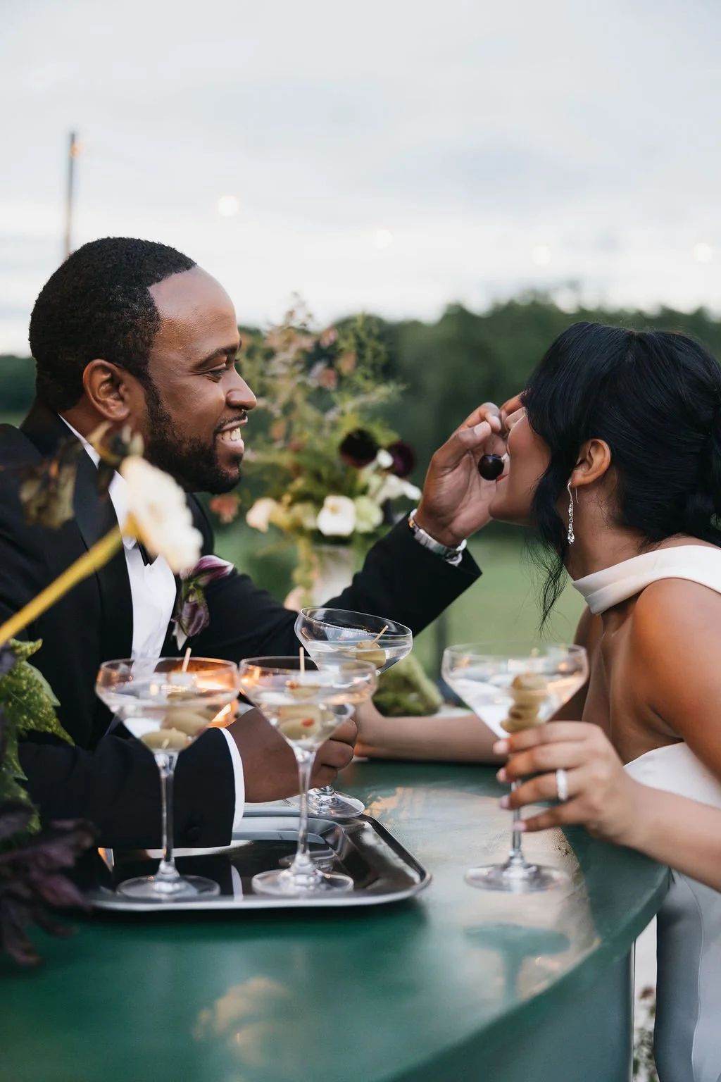 A man in a tuxedo is playfully feeding a woman at an outdoor wedding reception, with drinks and floral decorations on the table.