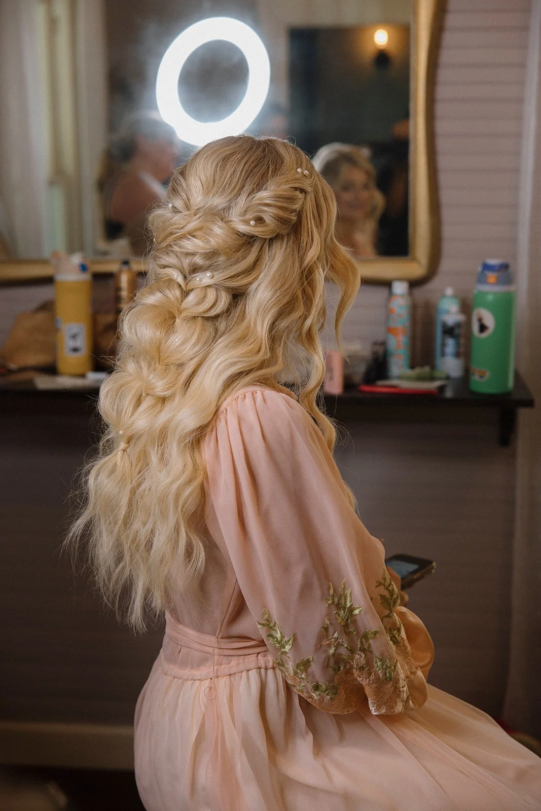 A woman with long, wavy blonde hair styled with small pearl accessories, sitting in front of a mirror in a dressing room, wearing a peach-colored dress with embroidered sleeves.