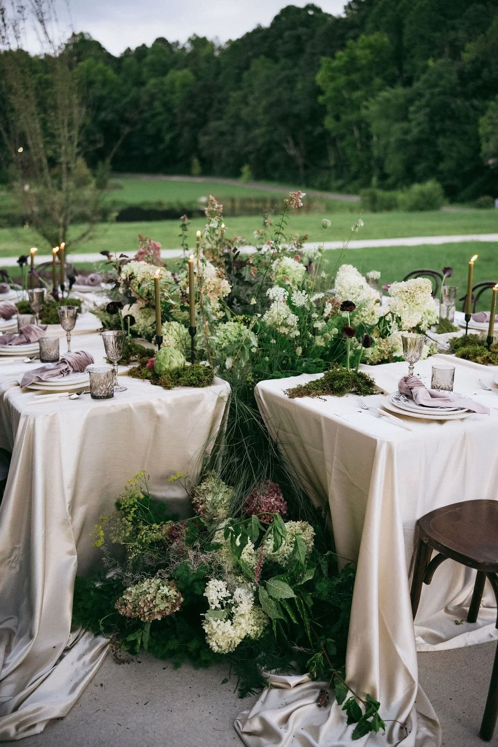 Elegant outdoor dining table decorated with candles and floral arrangements, set on white tablecloths, with a lush green landscape background during dusk.