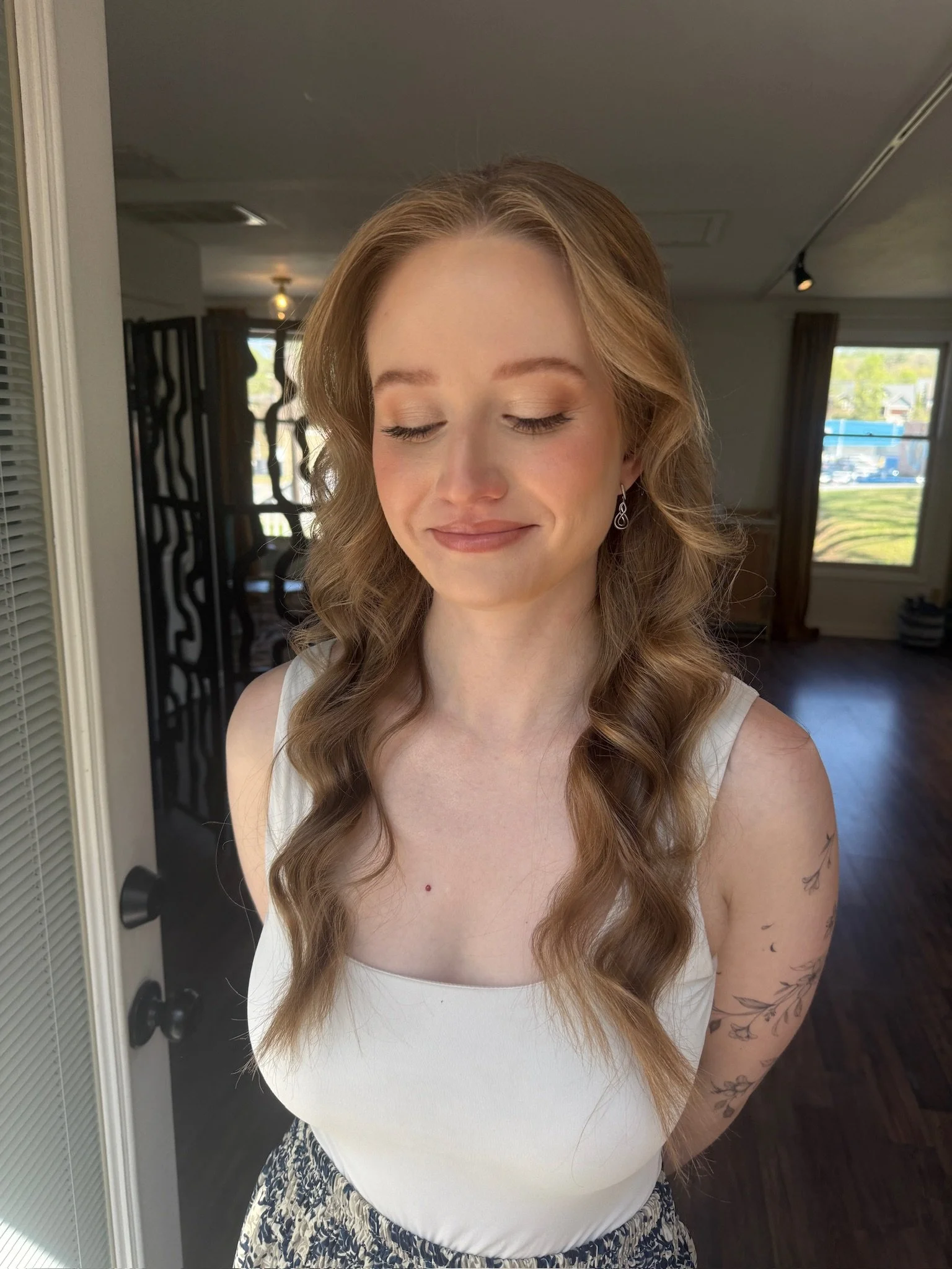 A woman with long, curly, reddish-brown hair, wearing a white sleeveless top, standing in a room with large windows and dark wooden flooring, her eyes closed and smiling gently.