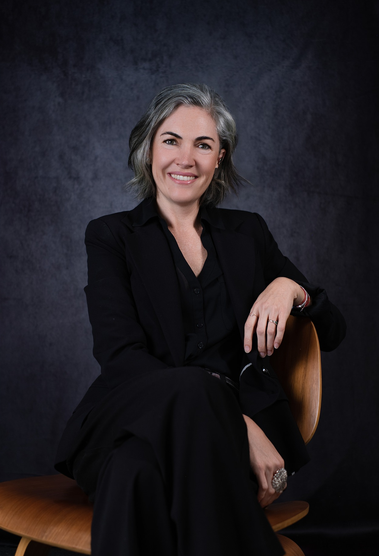 A woman with gray hair wearing a black blazer and black pants, sitting on a wooden chair with a dark textured background, smiling and looking at the camera.