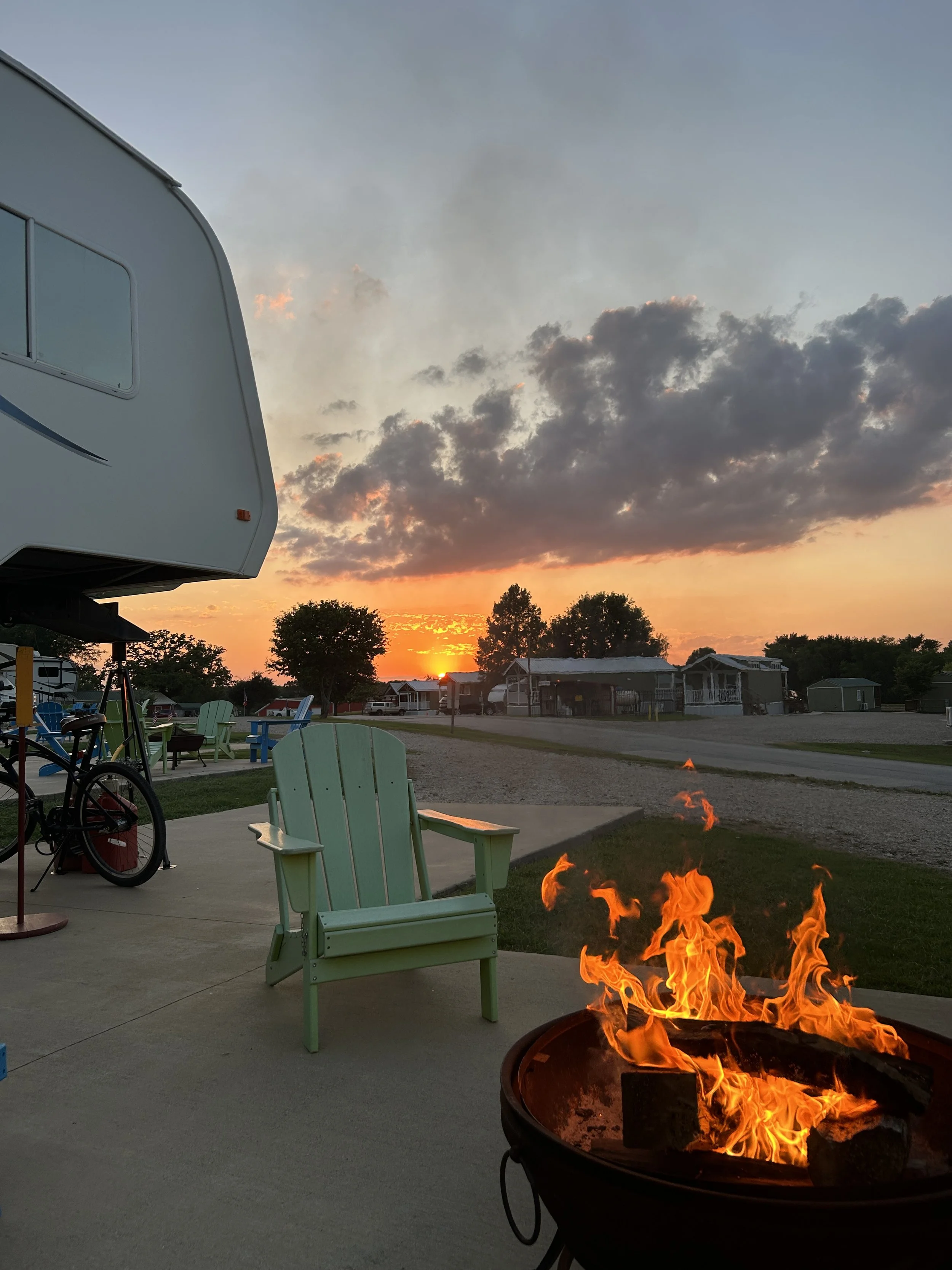 A fire pit with flames burning on a patio, green outdoor chairs, a camper trailer, trees, and houses in the background at sunset.