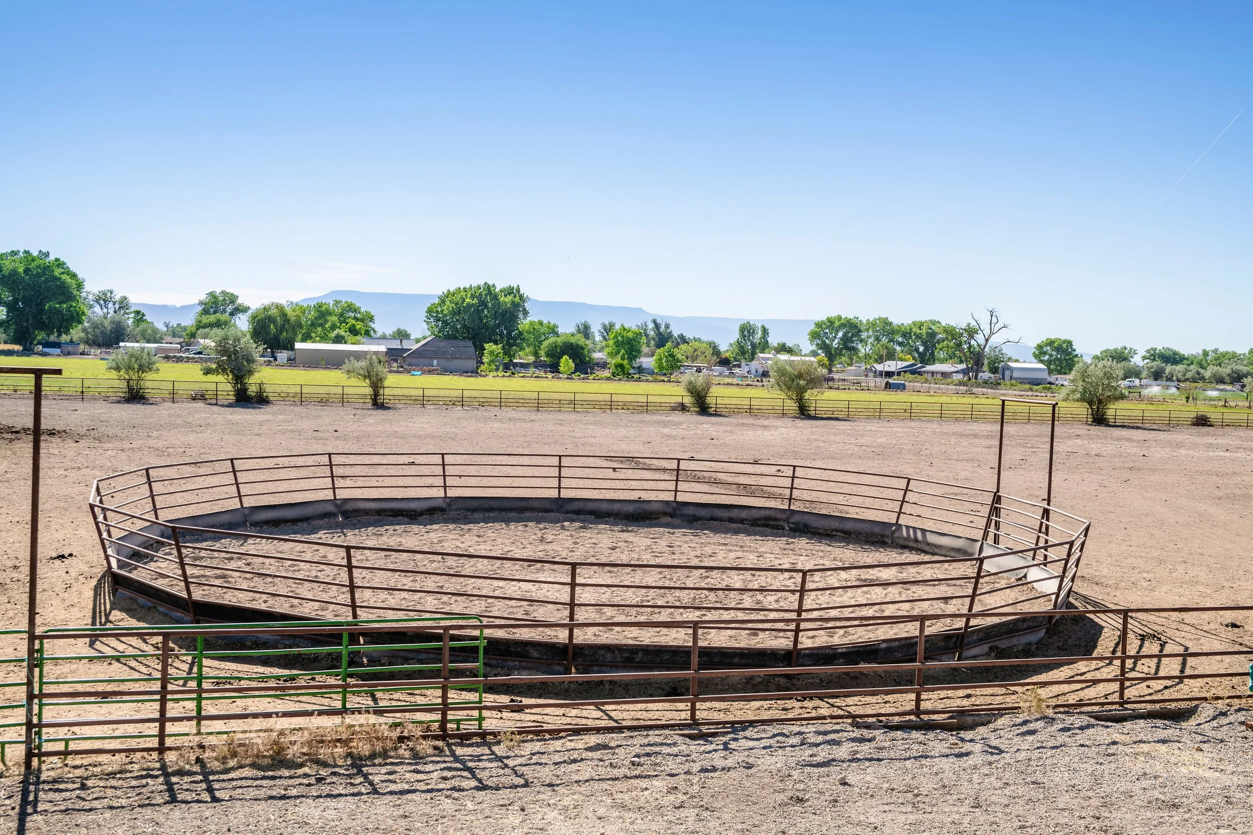 A circular animal enclosure with reddish-brown metal fencing on dusty ground, set against a backdrop of green trees, scattered buildings, and distant mountains under a clear blue sky.