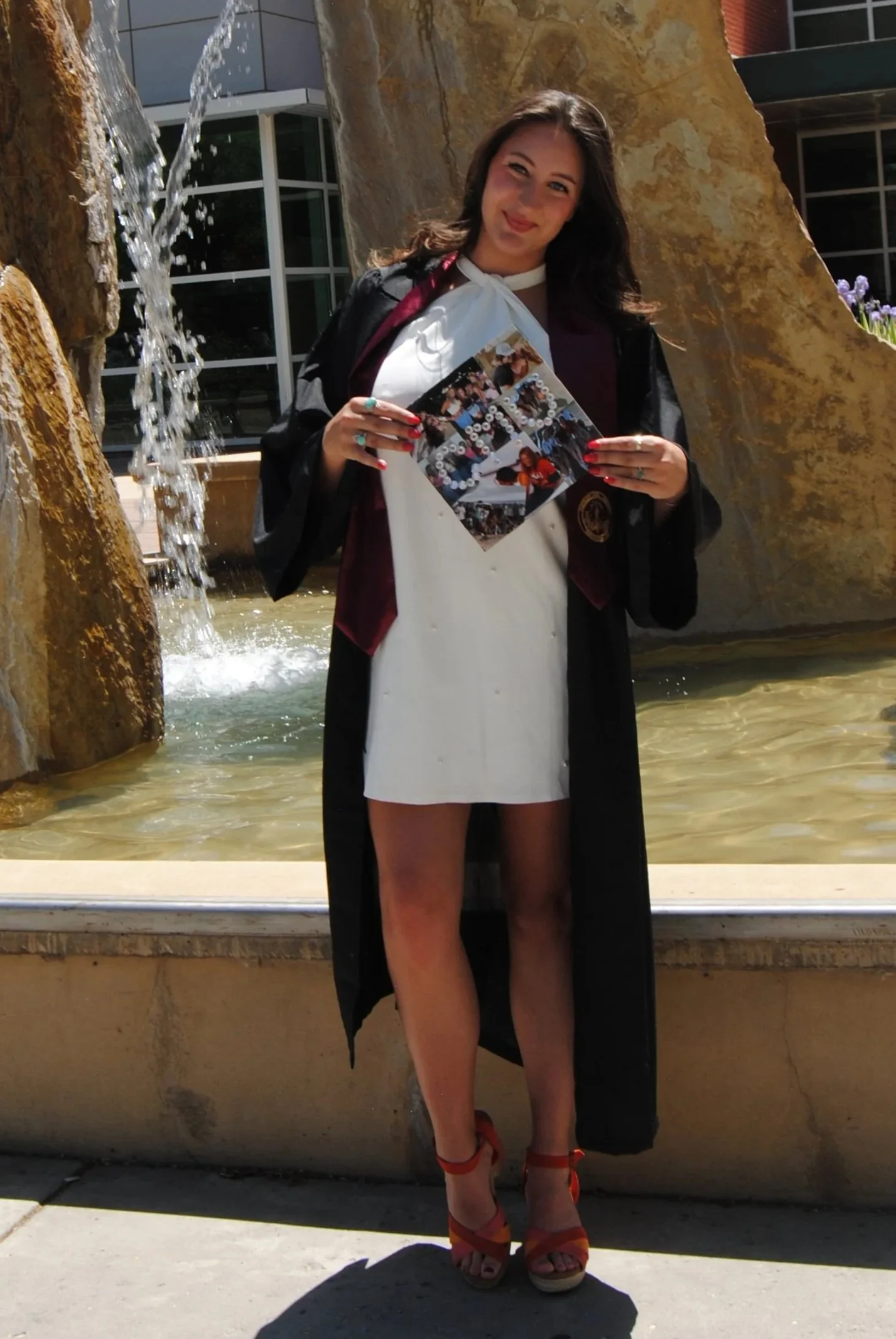 A young woman in a graduation gown and white dress, holding a photo collage, standing in front of a fountain with water cascading down rocks, smiling at the camera.
