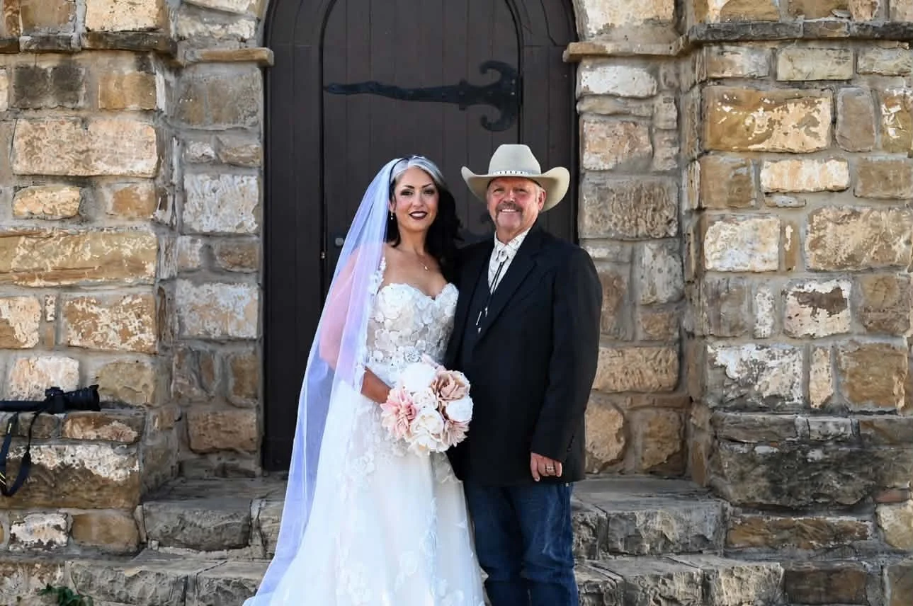 A bride and an older man, possibly her father, standing together in front of a stone building with a dark wooden door. The bride is wearing a white wedding dress with floral details, a sheer veil, and holding a bouquet of pale pink and white flowers. The man is dressed in a dark suit with a white shirt and a light-colored cowboy hat. Both are smiling.