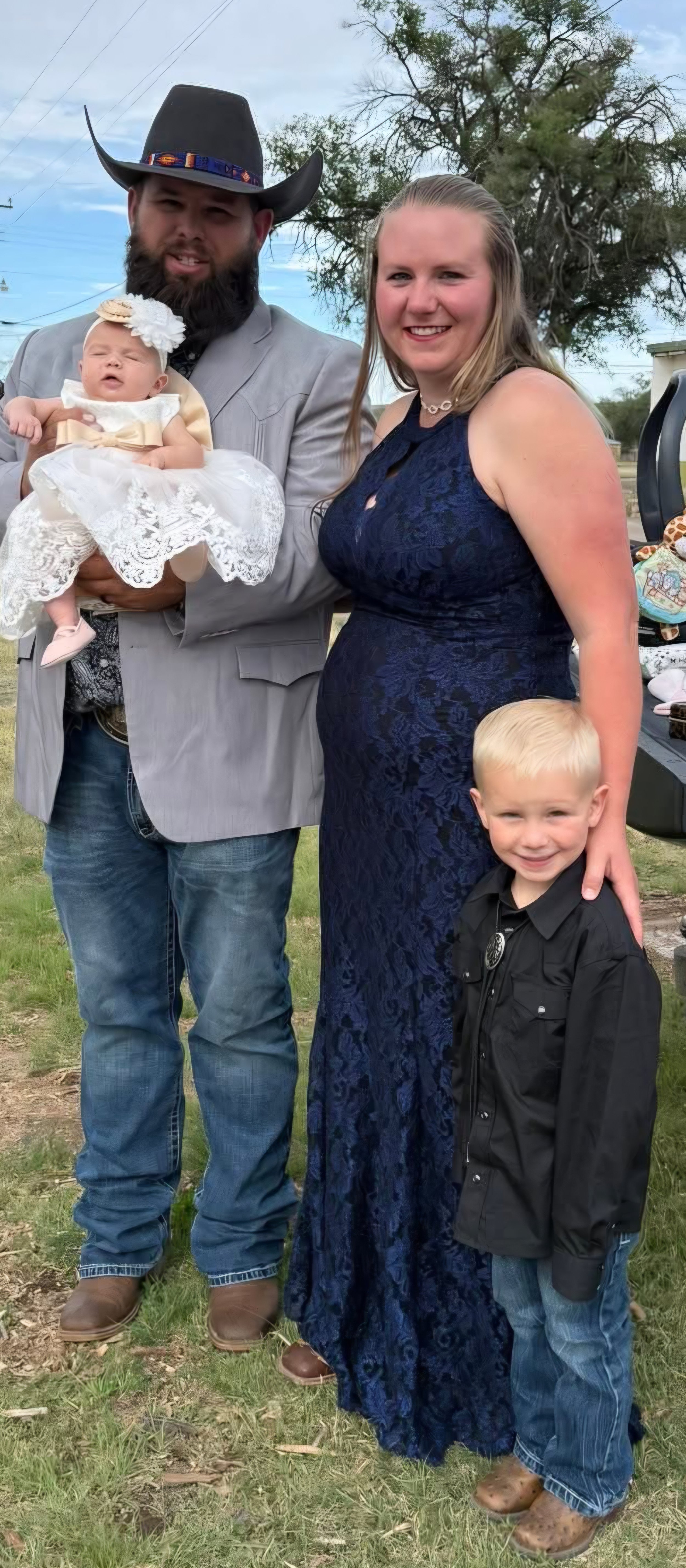 Family photo outdoors with a man in a cowboy hat, woman in a blue dress, a baby girl in a white dress, and a young boy in black shirt and jeans, smiling.