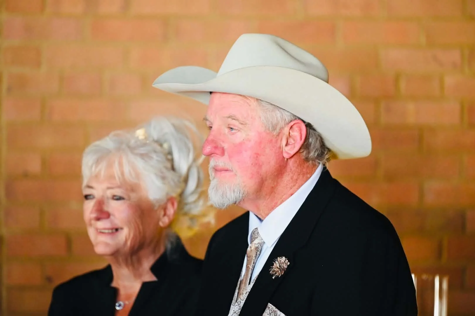 An elderly man in a black suit, white shirt, and patterned tie, wearing a large white cowboy hat with a feather pin, is sitting next to an elderly woman with gray hair and a black outfit, against a brick wall background.