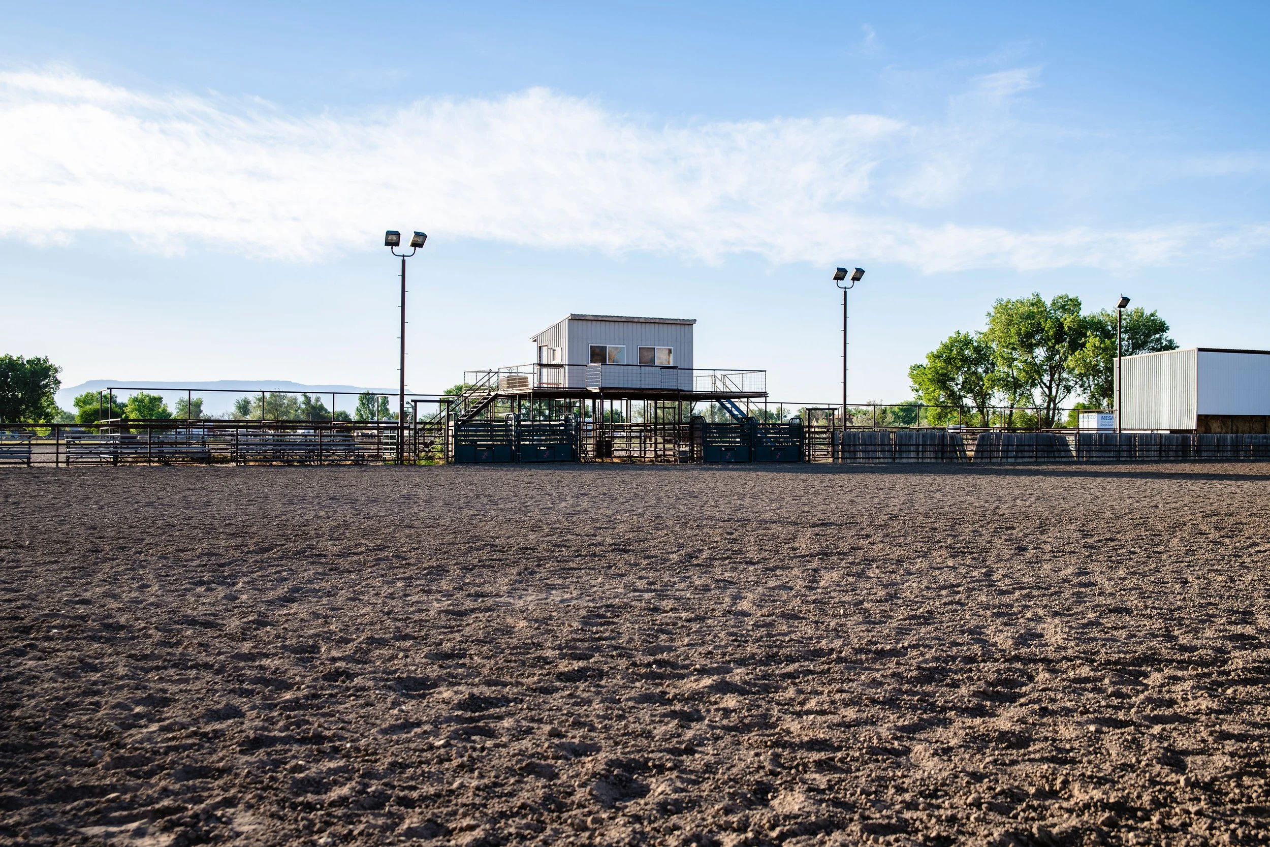 An empty dirt rodeo arena with a small elevated building in the center, surrounded by fencing. There are four tall light poles and some trees and blue mountains in the background under a partly cloudy sky.