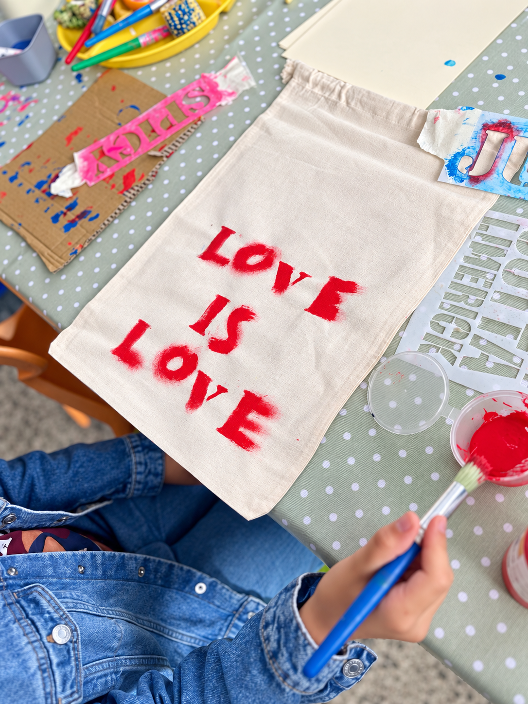 A person painting the words 'Love is Love' in red on a beige tote bag at a craft table with art supplies, stencil letters, and paints.