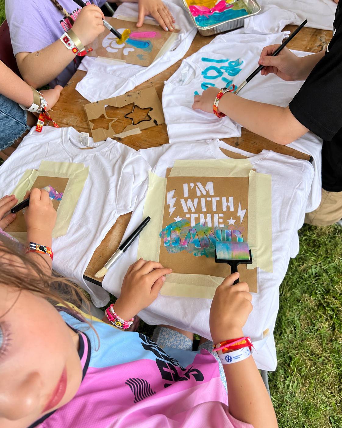 Children creating colorful art on T-shirts at an outdoor craft activity table. The T-shirts read 'I'M WITH THE BAND' with tie-dye and painted designs.