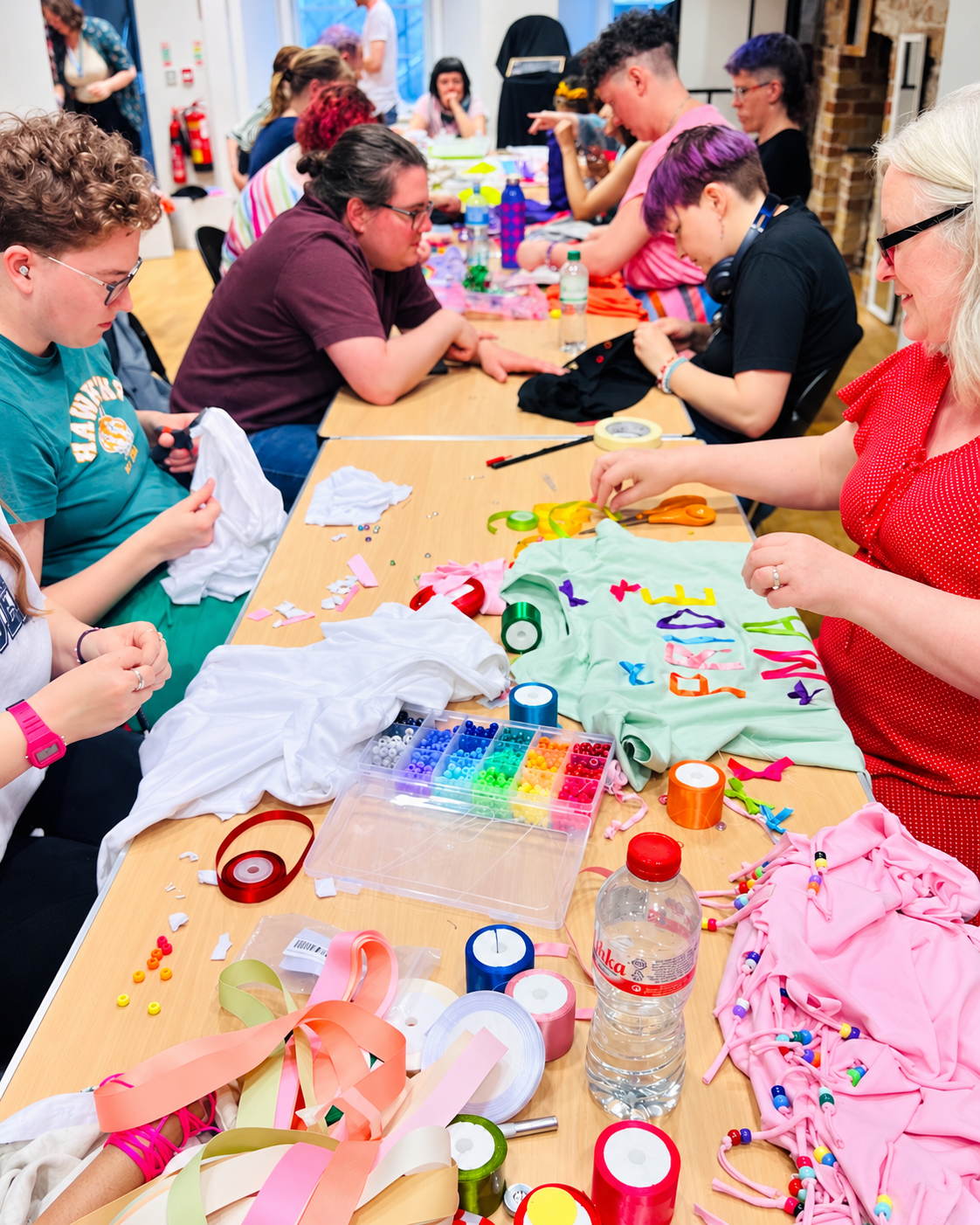 People sitting at a long table engaging in crafts and decorations, with various fabrics, ribbons, beads, and craft supplies spread on the table.