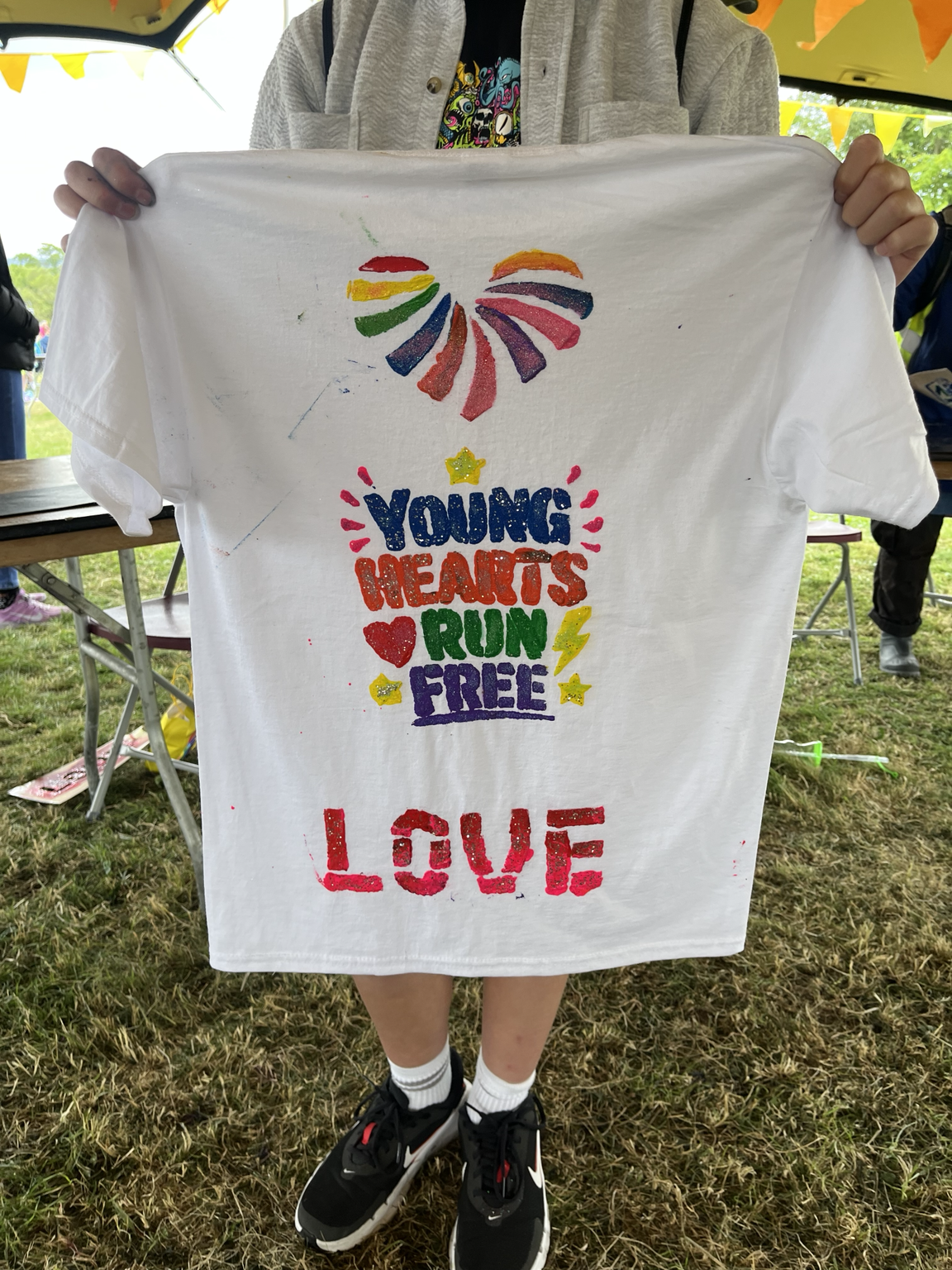 A person is holding a white t-shirt with colorful text and images. The shirt features a rainbow-colored heart at the top, the words 'YOUNG HEARTS RUN FREE' in multicolored bold letters, a small yellow star above the text, a red heart, yellow lightning bolt, and the word 'LOVE' at the bottom in red. The person's face is not visible, and they are standing outdoors on grass under a yellow canopy with bunting. They are wearing black sneakers, white socks, jeans, and a jacket.